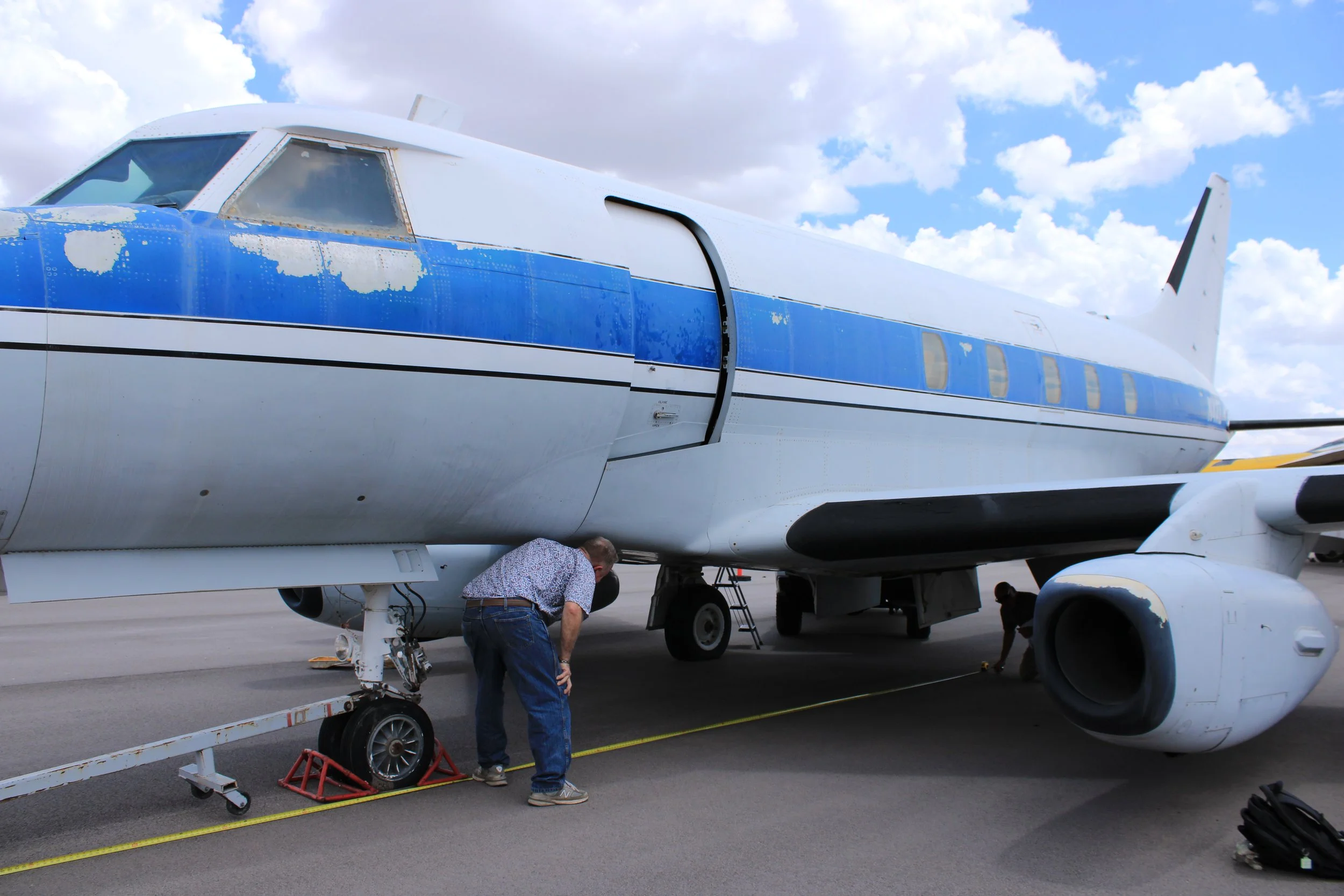 A person working on a white and blue private jet aircraft on the tarmac, with a cloudy sky overhead.