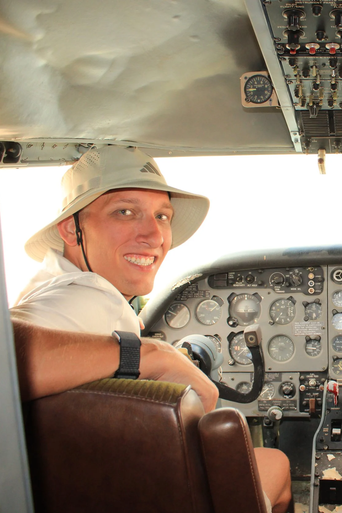 A man sitting in the cockpit of an aircraft, wearing a beige sun hat and a white shirt, smiling at the camera with the airplane control panel and instruments visible around him.