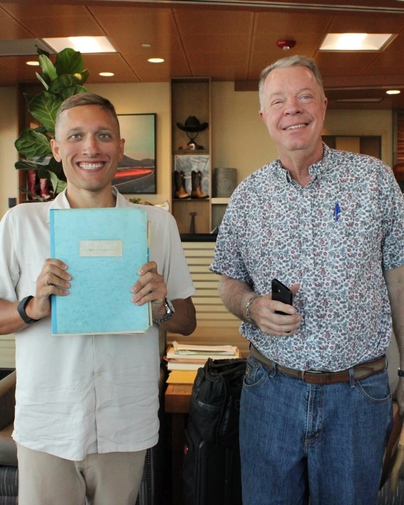 Two smiling men standing indoors, one holding a blue folder and the other holding a smartphone, with books and decor in the background.