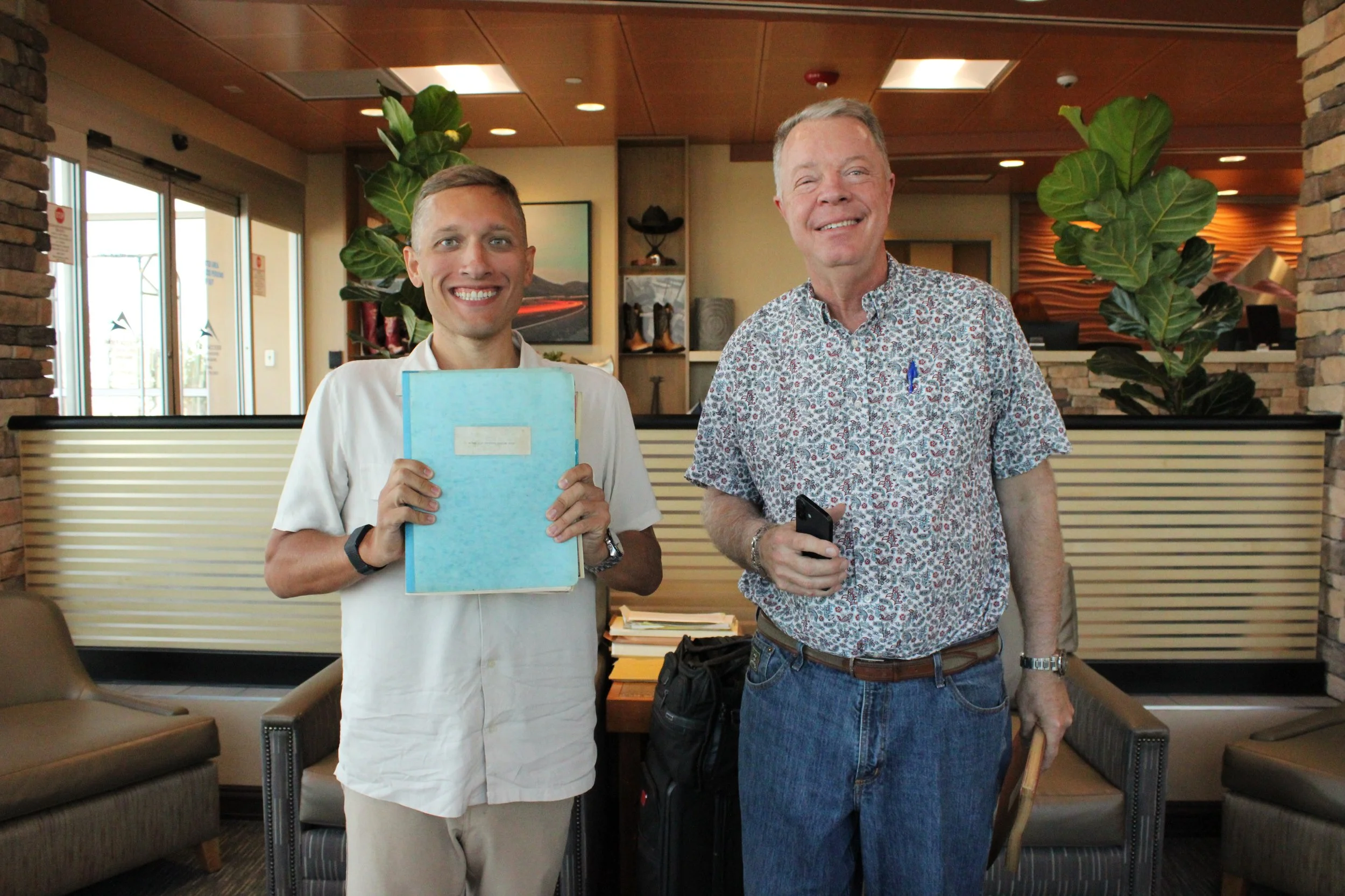 Two men standing inside a modern office or hotel lobby, smiling at the camera. The man on the left is holding a blue folder and wearing a white shirt, while the man on the right is dressed in a patterned shirt and jeans, holding a smartphone. There are indoor plants and artwork in the background.