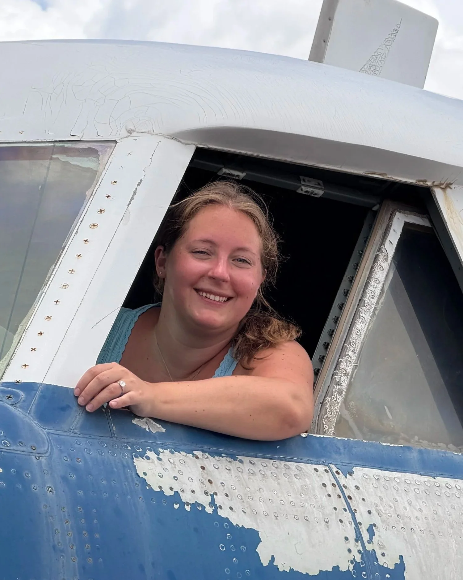 A smiling woman with light skin and brown hair leaning out of the cockpit of a vintage airplane, with an aged and weathered blue and silver exterior.