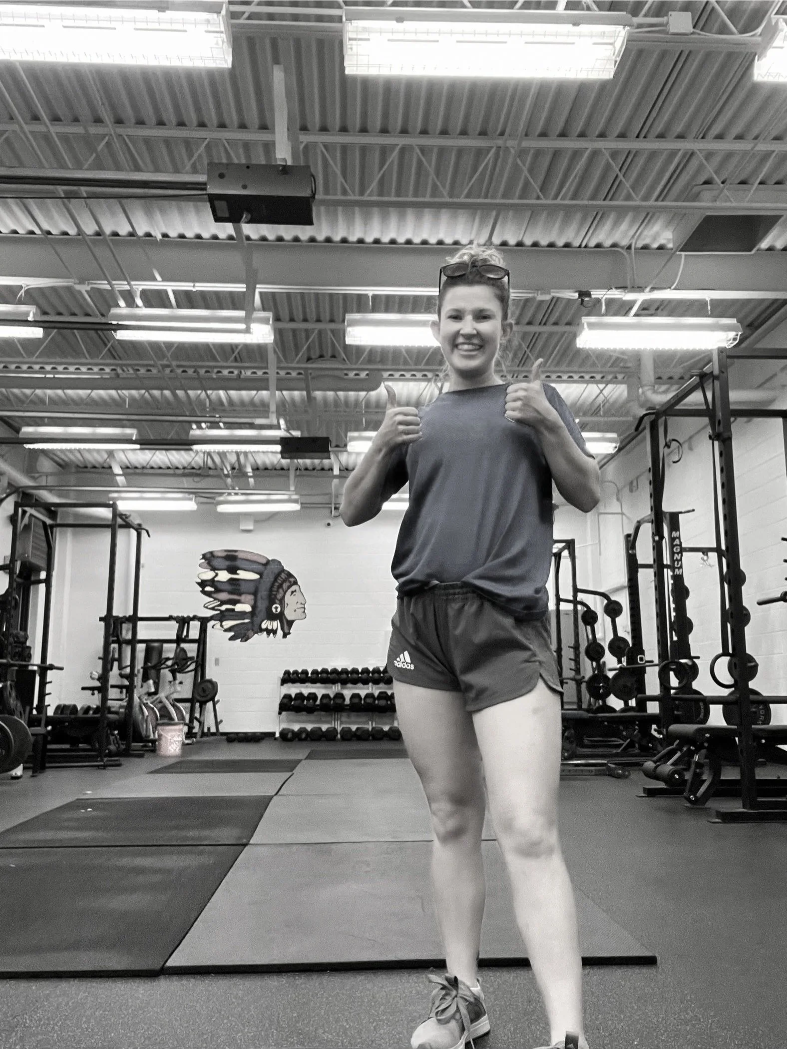 A young woman in athletic clothing giving a thumbs-up in a gym with weights and equipment in the background.