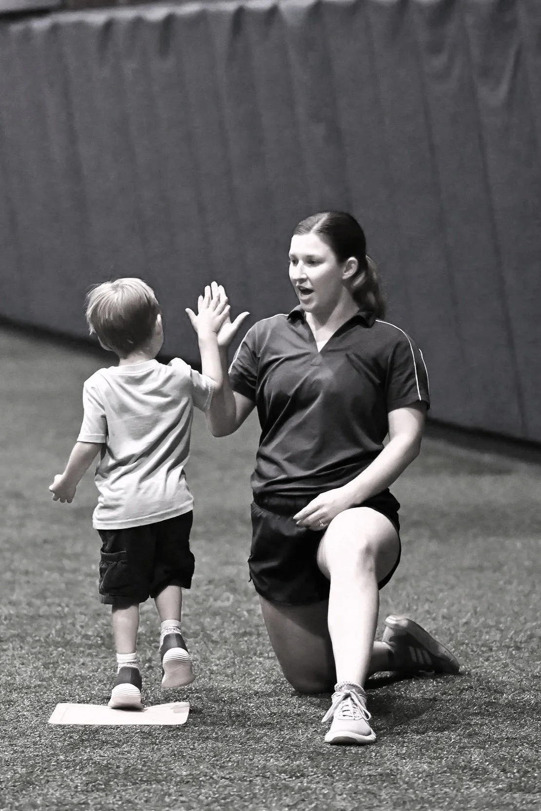 A woman kneeling on one knee giving a high five to a young boy who is running toward her, on an indoor sports field.