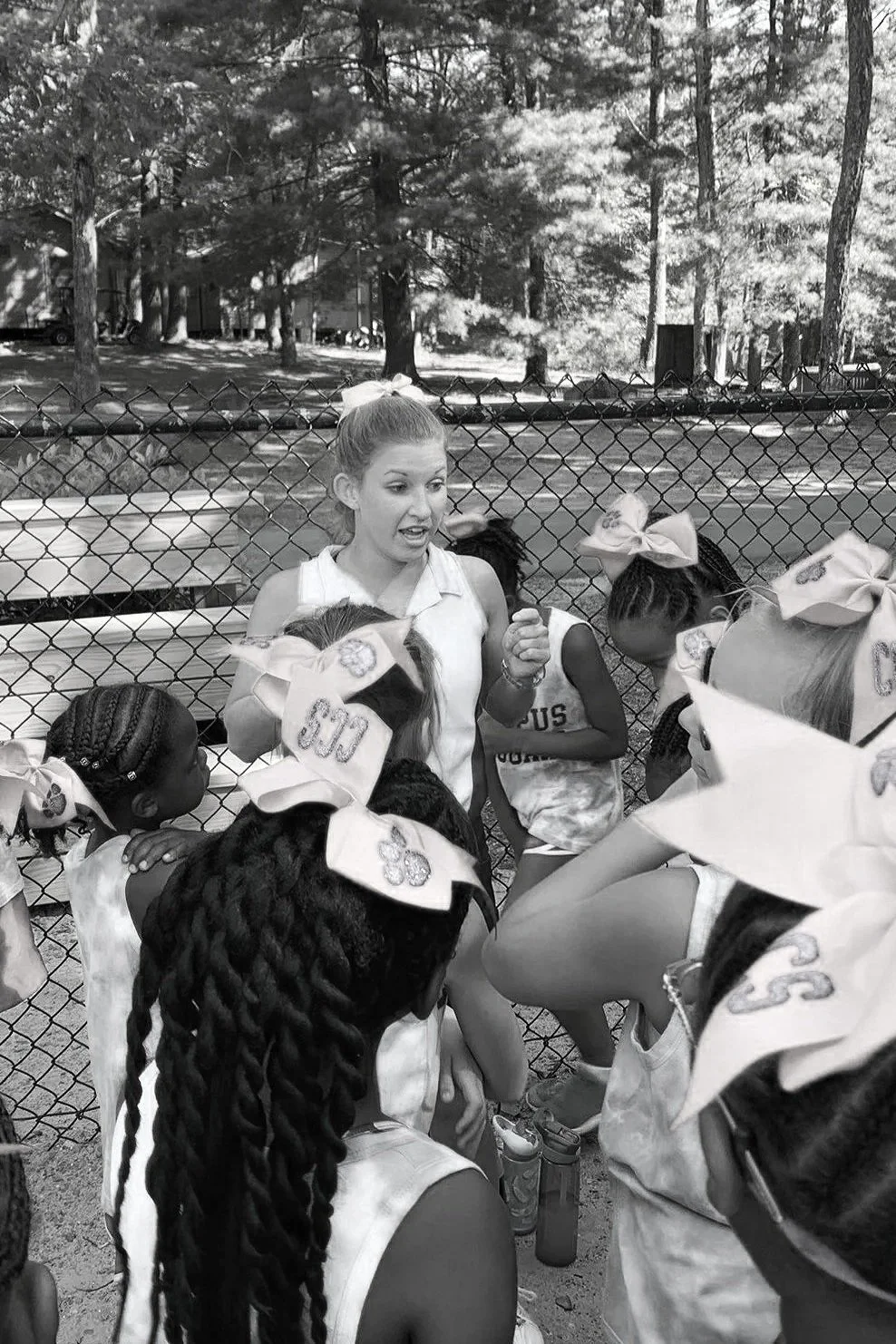 A coach talking to young female athletes during a break or discussion outdoors, with trees and a fence in the background.