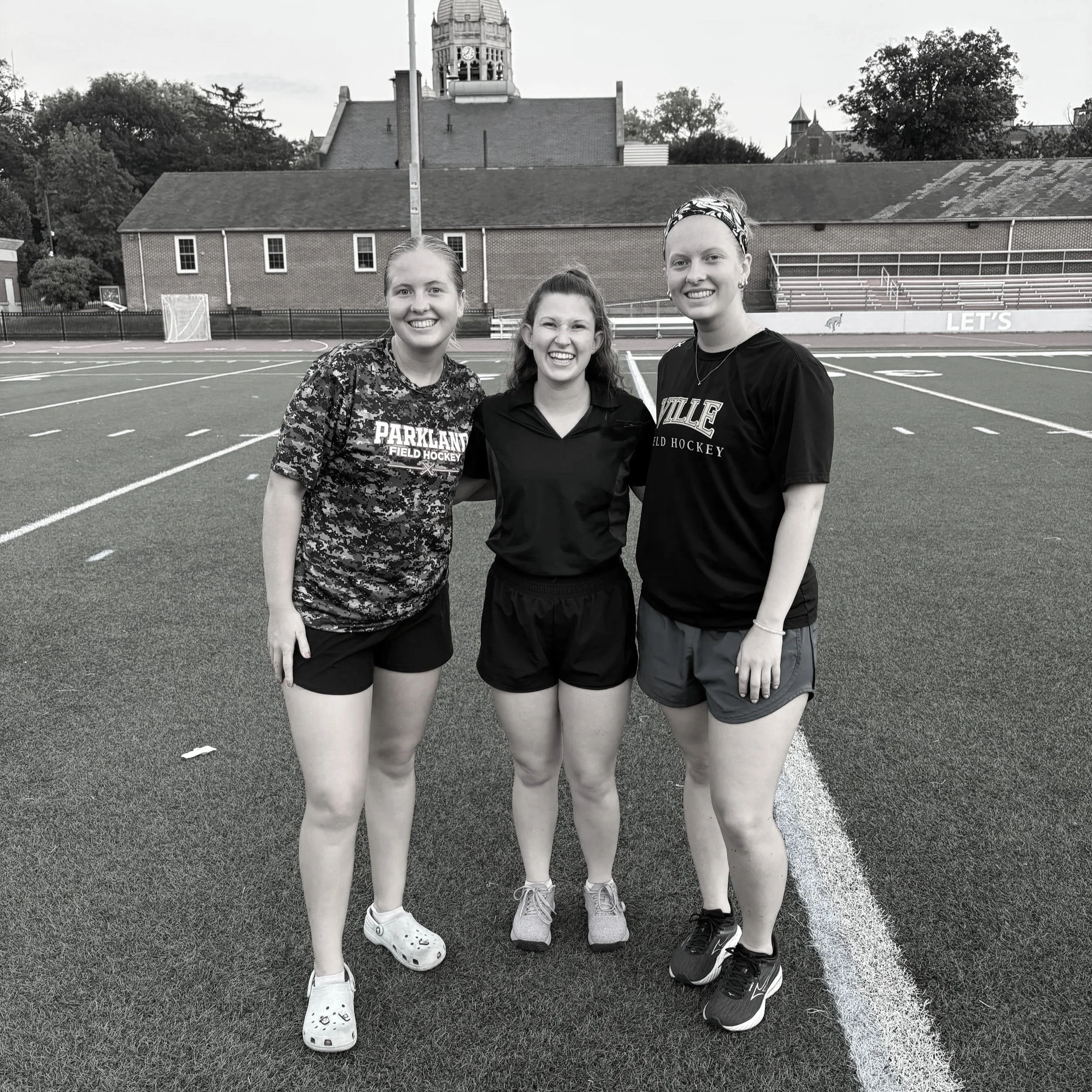Three young women standing on a sports field, smiling, with a school building and trees in the background.