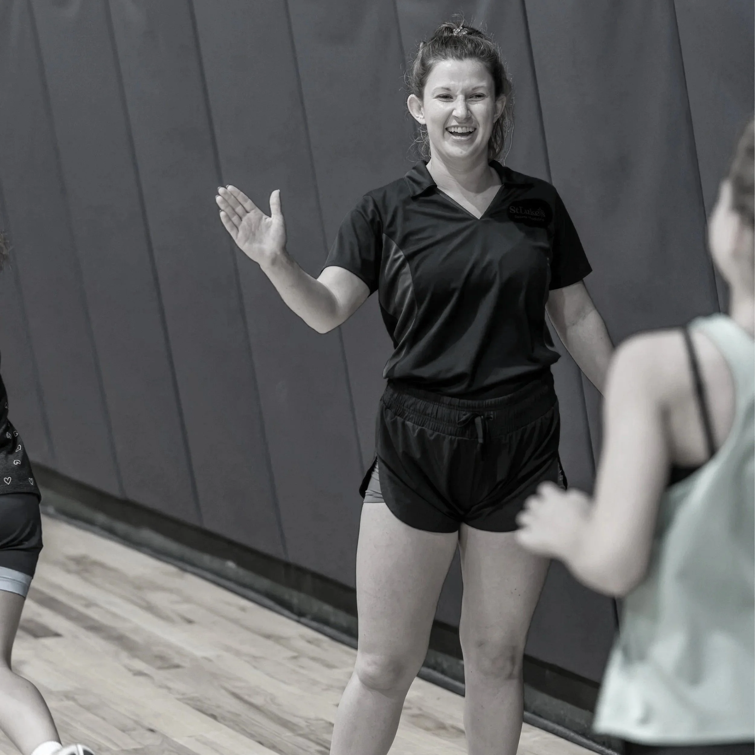 A woman with curly hair in a bun, dressed in a black athletic shirt and shorts, high-fiving an athlete and smiling in what appears to be a gym or fitness studio. She is standing on a wooden floor in front of a dark wall.