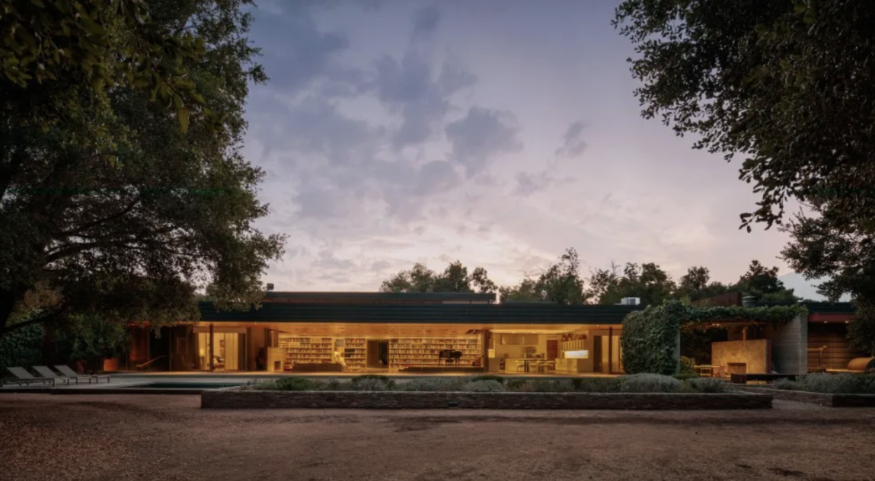 A low, glass-walled house in the Ojai Valley lit from within at dusk, framed by mature oaks with the dark treeline behind it.
