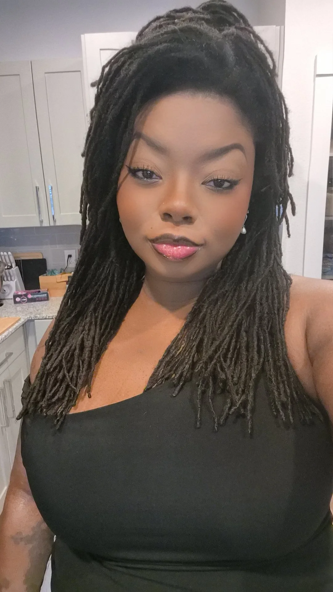 A woman with long dreadlocks and makeup taking a selfie in a kitchen.