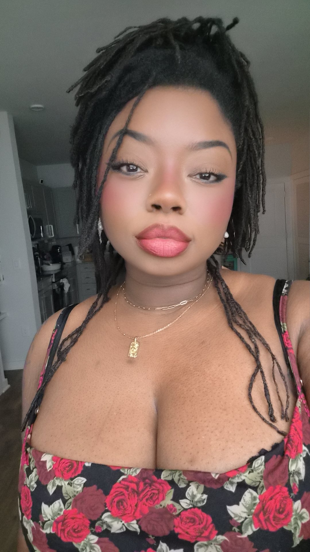 A woman with dreadlocks wearing a floral dress, multiple necklaces, and earrings, taking a selfie in a room with a kitchen in the background.
