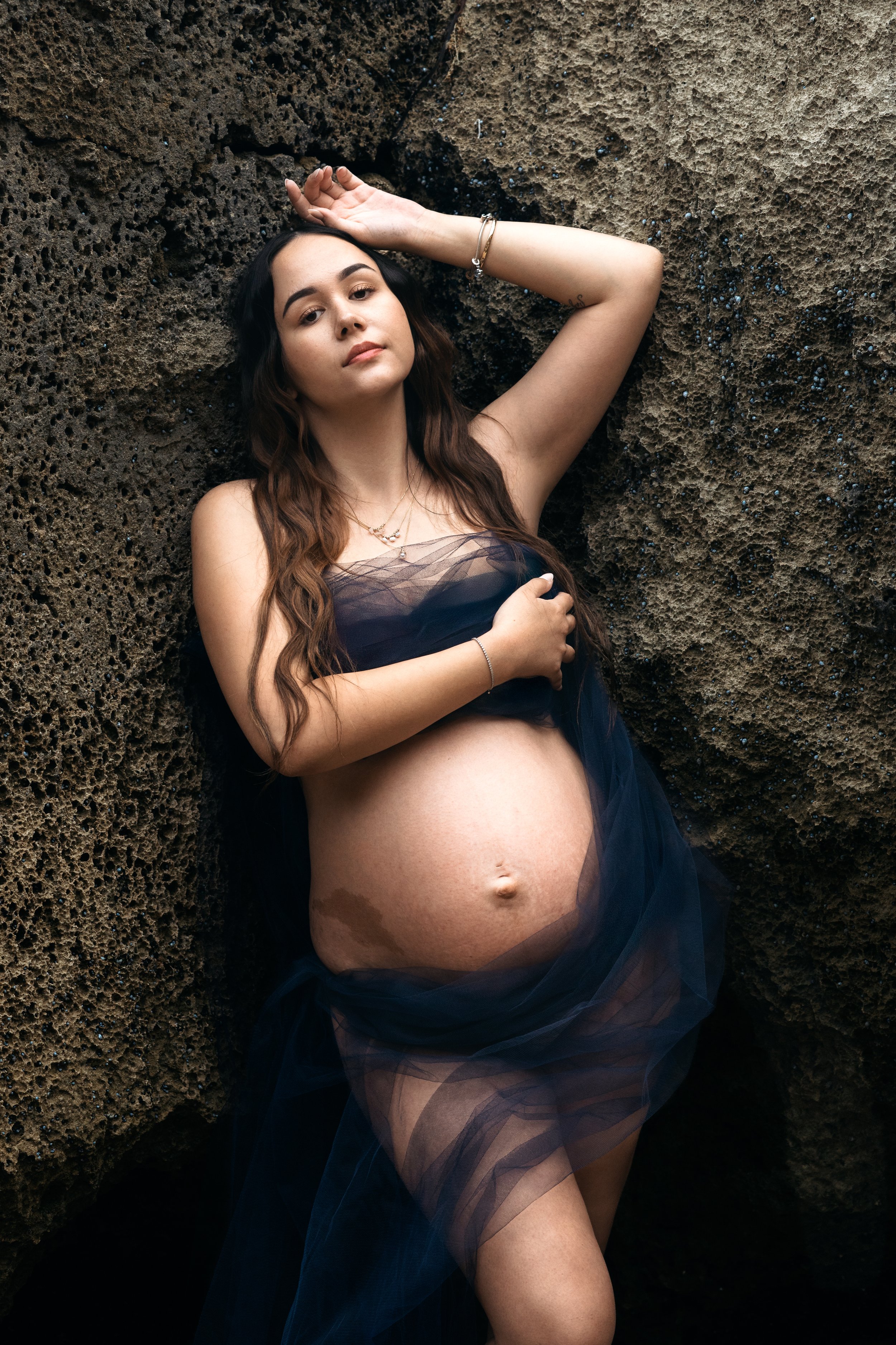 A pregnant woman with long brown hair poses against a textured volcanic rock surface. She wears a sheer navy blue fabric draped over her body, revealing her pregnant belly. She has jewelry including necklaces and bracelets, with her arms positioned t