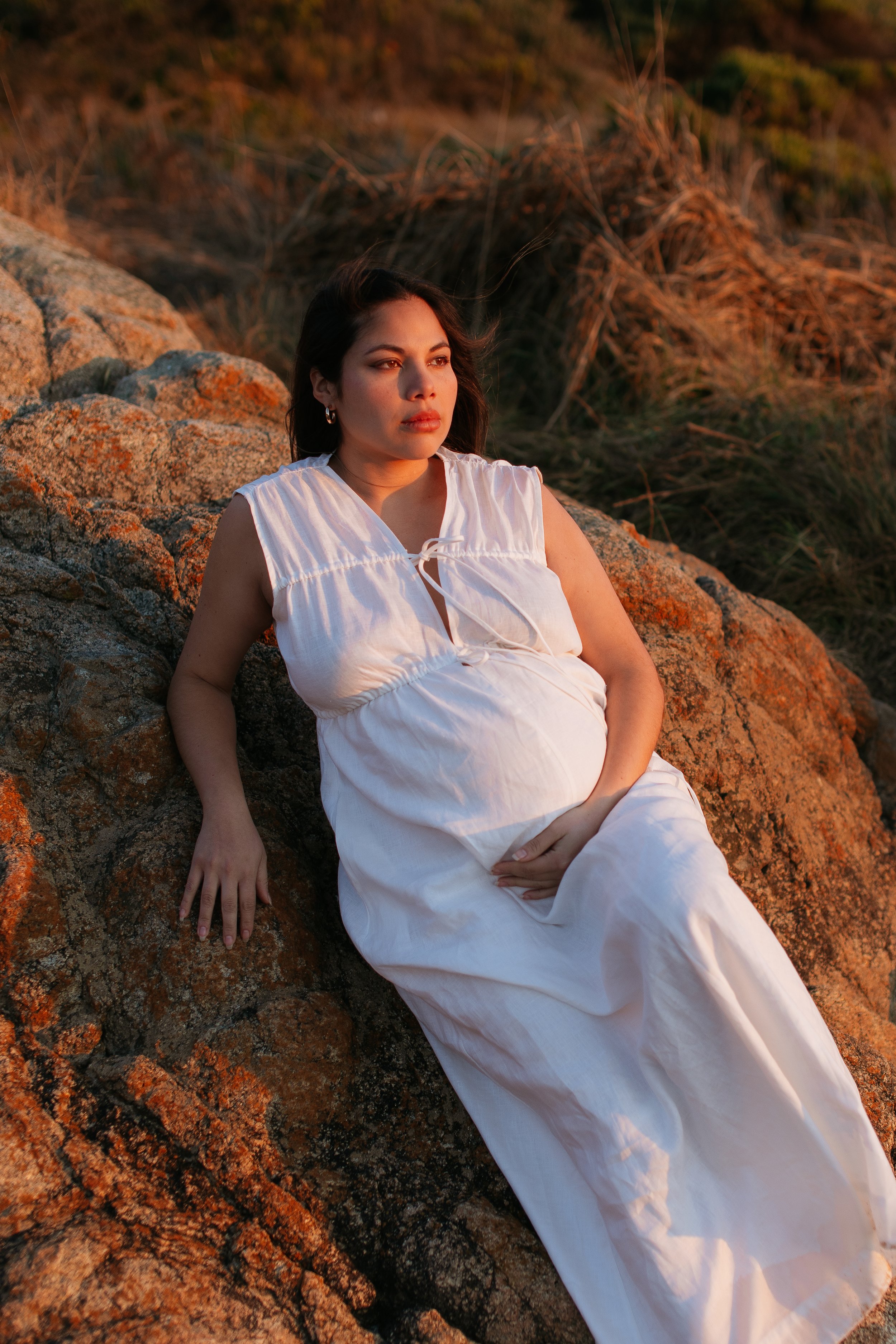A woman in a white dress sitting on rocks outdoors during sunset, with her hand on her lower stomach and looking thoughtfully into the distance.
