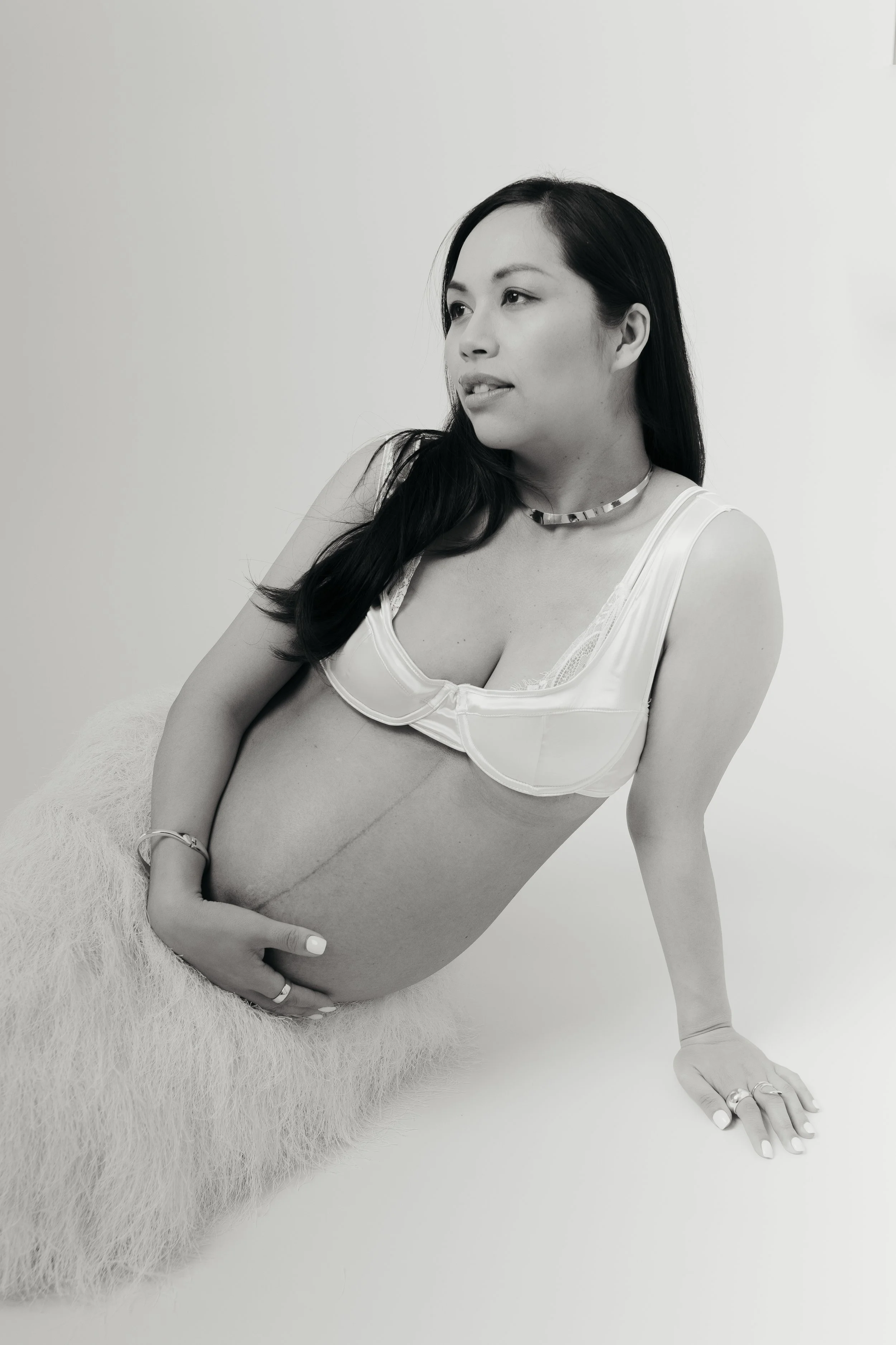 Black and white photograph of a pregnant woman sitting on a soft surface, resting her hand on her belly, looking to the side with a slight smile, wearing a white top and jewelry.