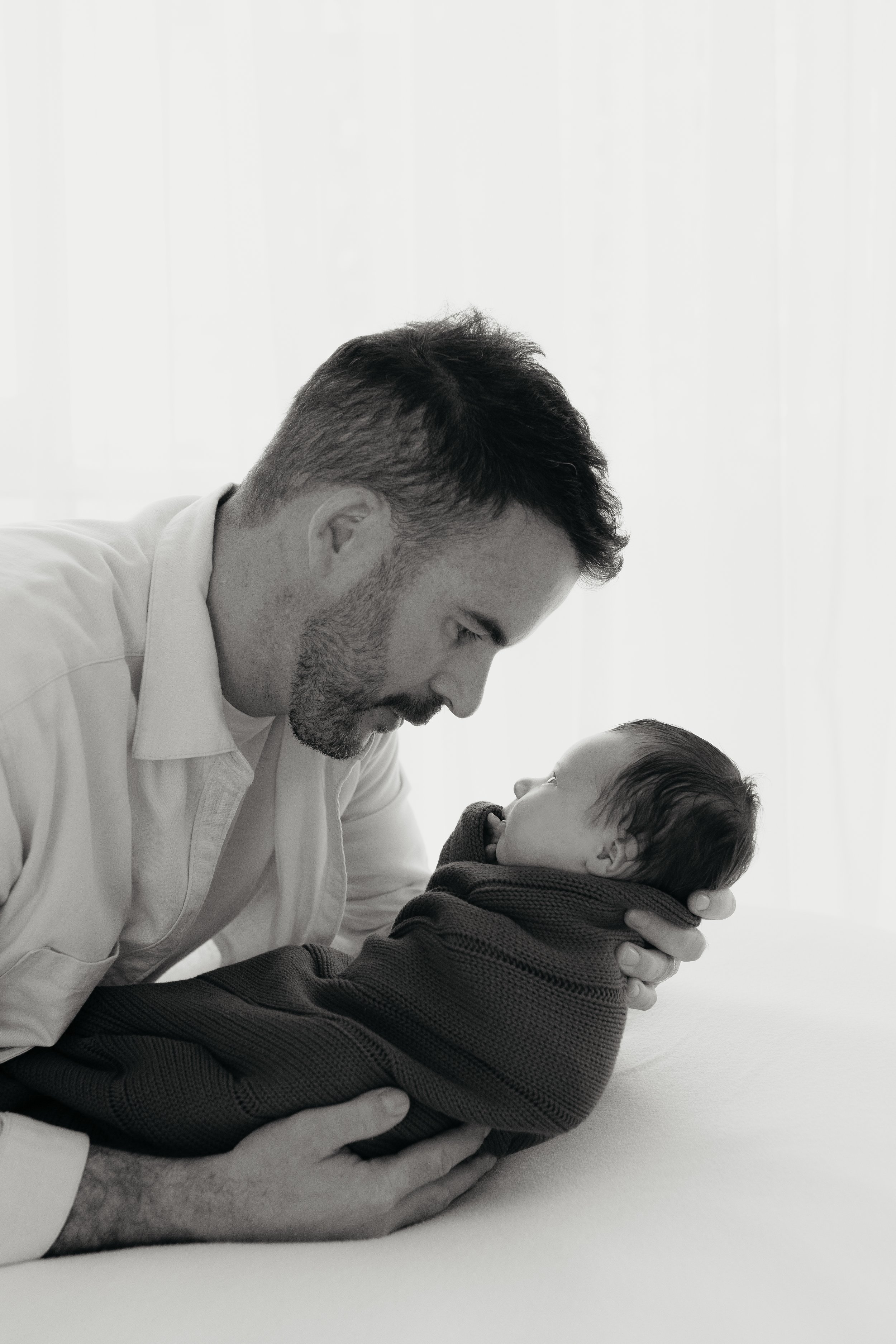 A man with dark hair and a beard gently holding a young child wrapped in a dark hoodie and blanket, looking into each other's eyes in a softly lit, minimalist room.