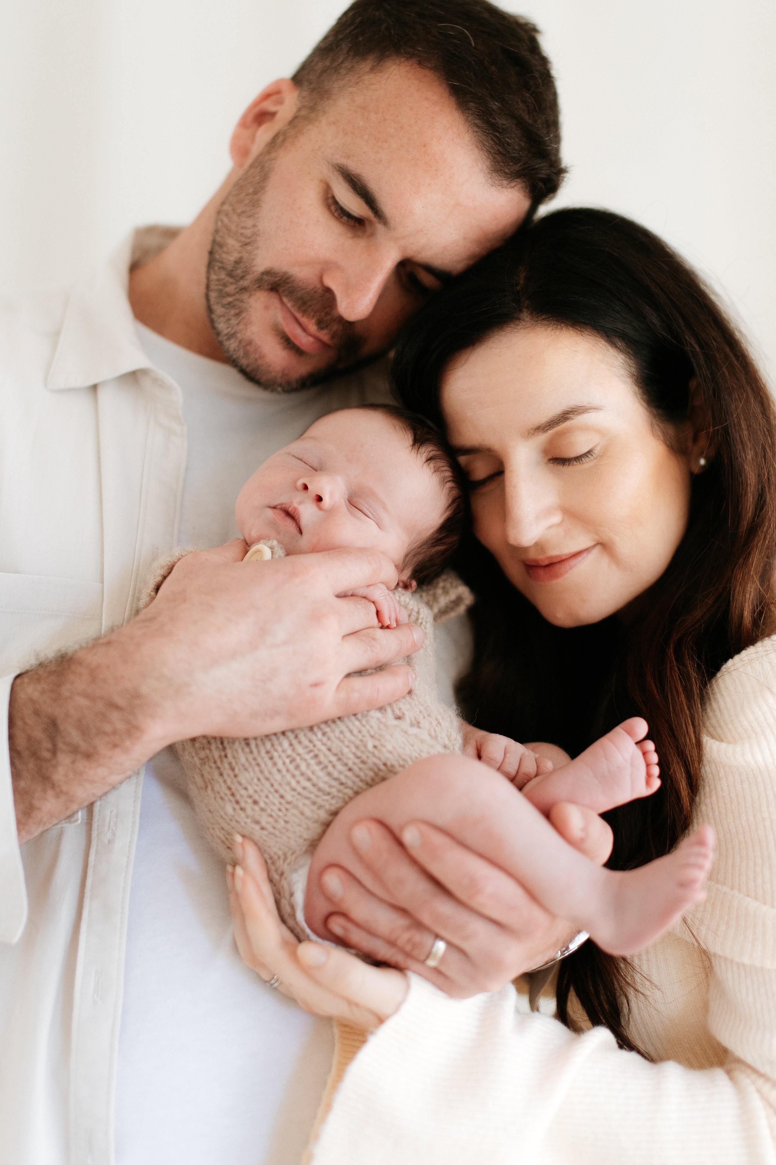 A man and woman lovingly holding a sleeping baby, their faces close together with gentle smiles.