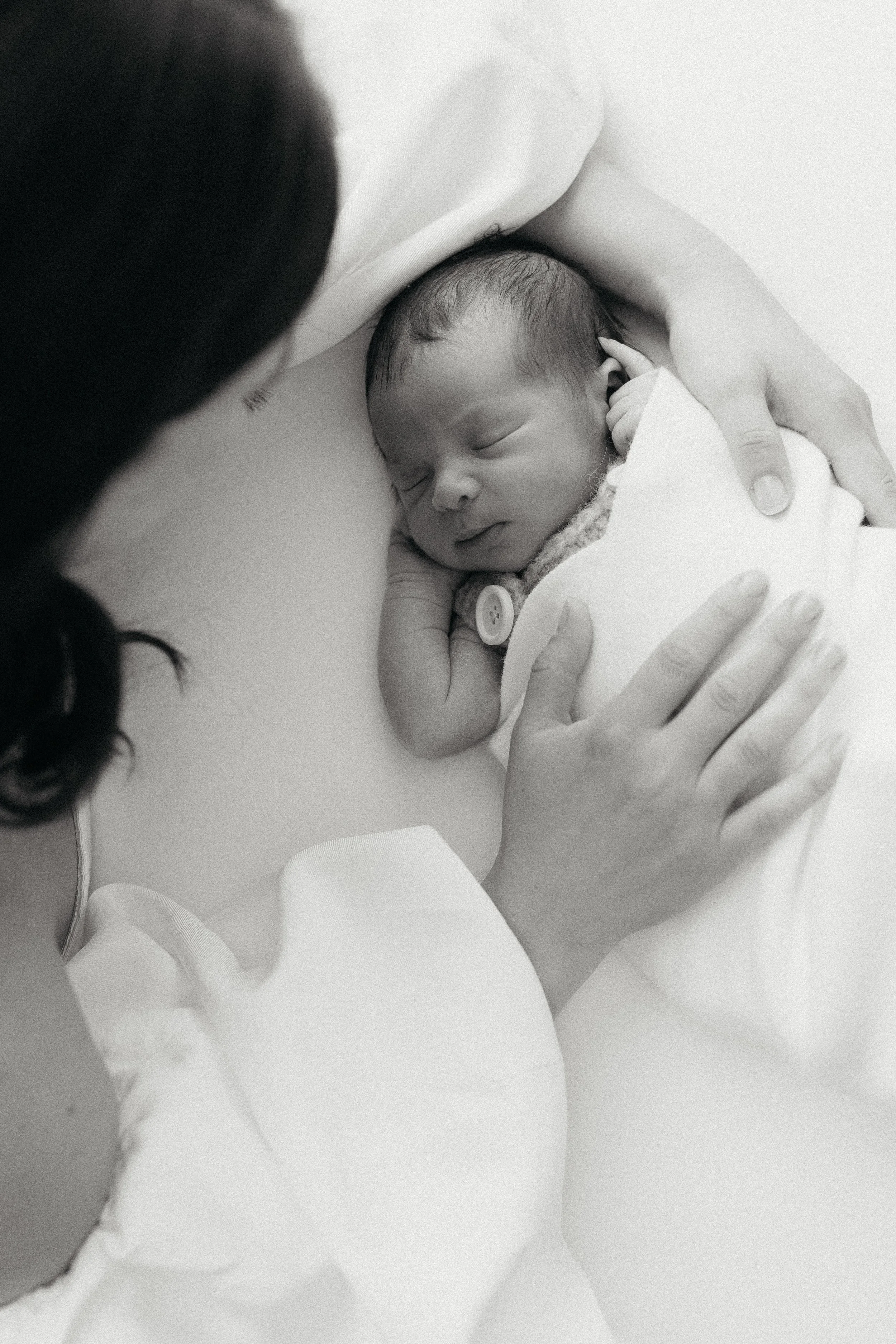 A person with dark hair holds a newborn baby close to their chest, both lying on a white surface. The baby is peacefully sleeping with a pacifier in their mouth and a tiny hand near their face.