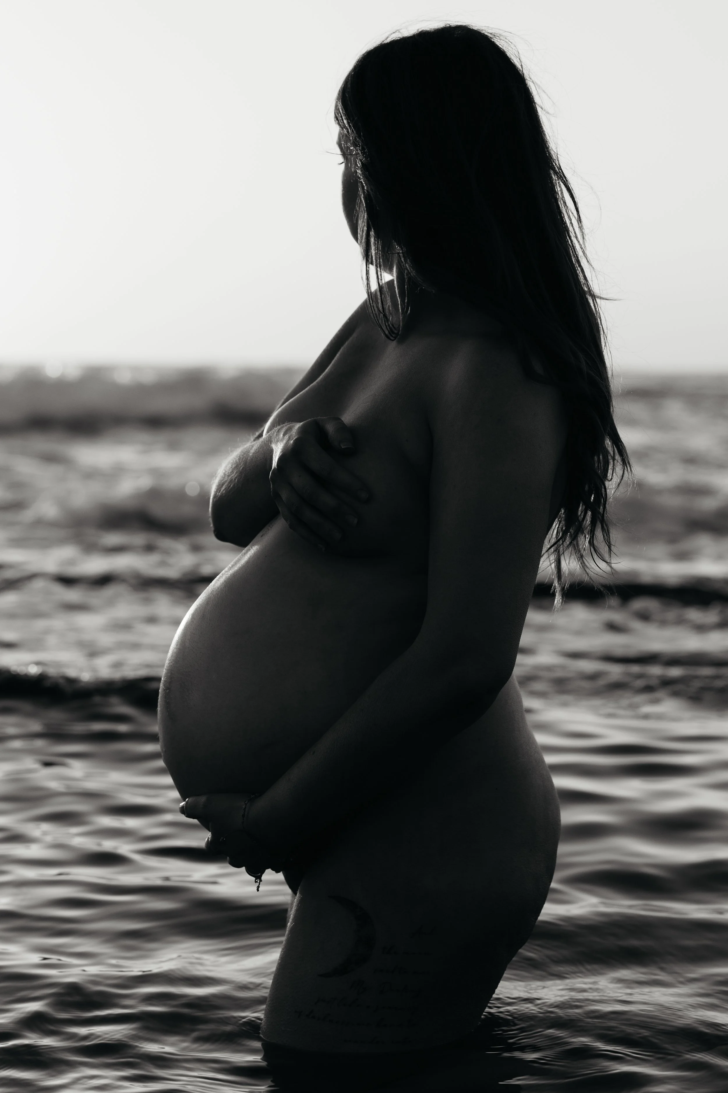 Silhouette of a pregnant woman standing in the ocean with arms crossed, captured in black and white.