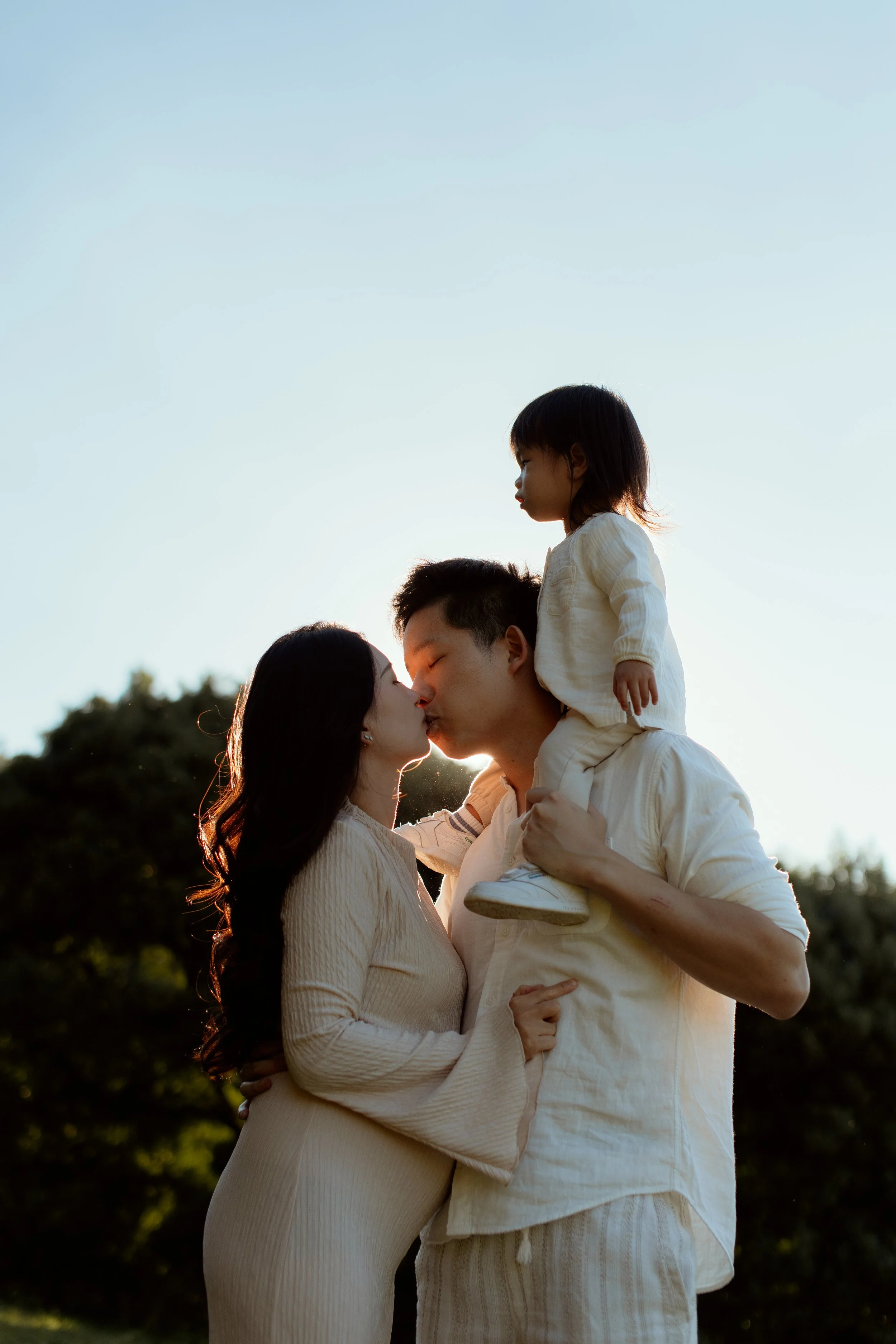 A family of three, a man, a woman, and a young girl, standing outdoors during sunset. The man is holding the girl on his shoulders, and they are about to kiss while the woman looks up at them with affection. The background features trees and a clear 