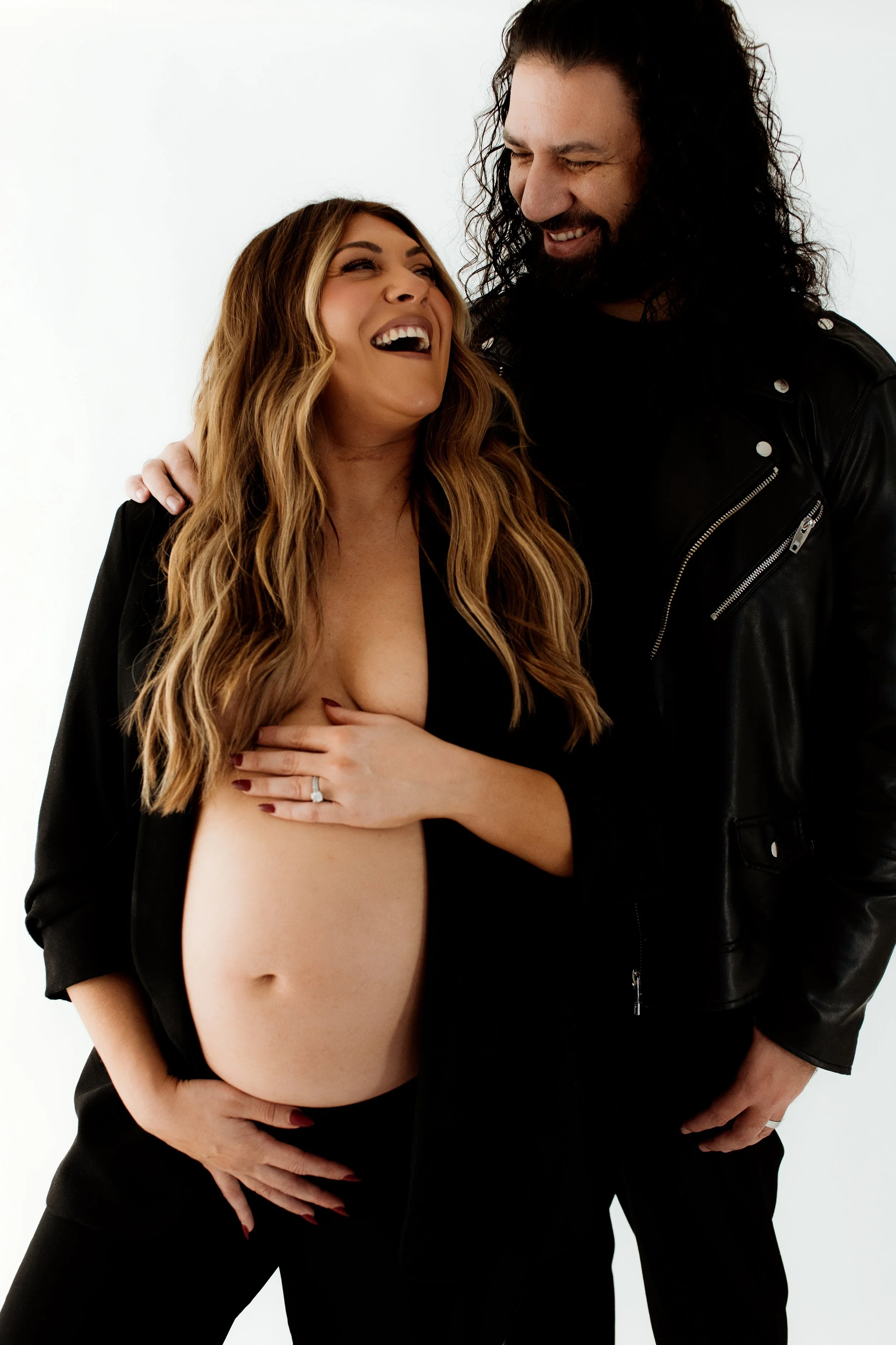 A woman with long wavy hair and a man with long curly hair, both laughing and smiling, stand close together against a plain white background. The woman is partially undressed, covering her chest with one hand, and wearing a black jacket. The man is w