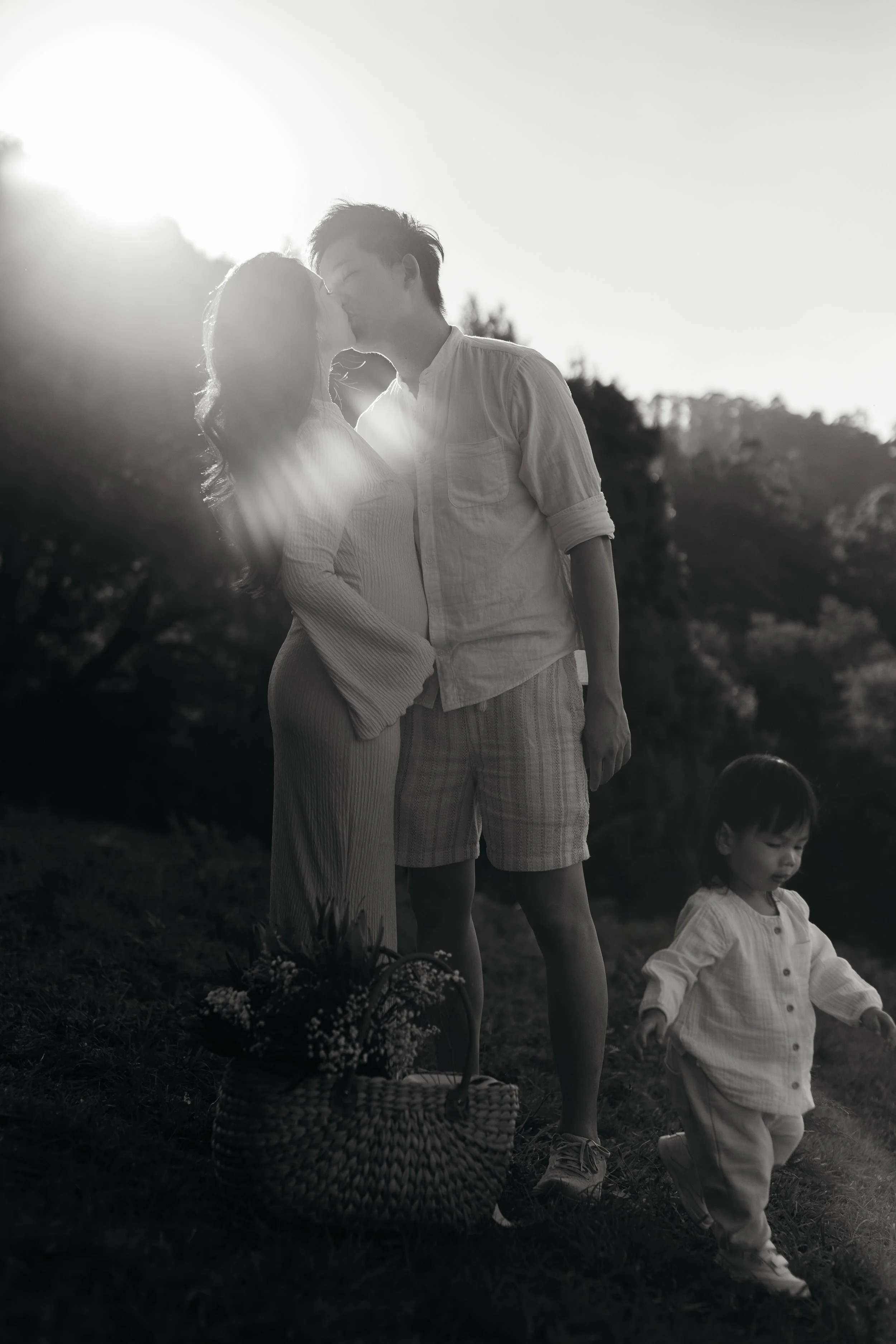 A black and white photo of a couple sharing a kiss outdoors with the sun setting behind them, and two young children walking nearby.