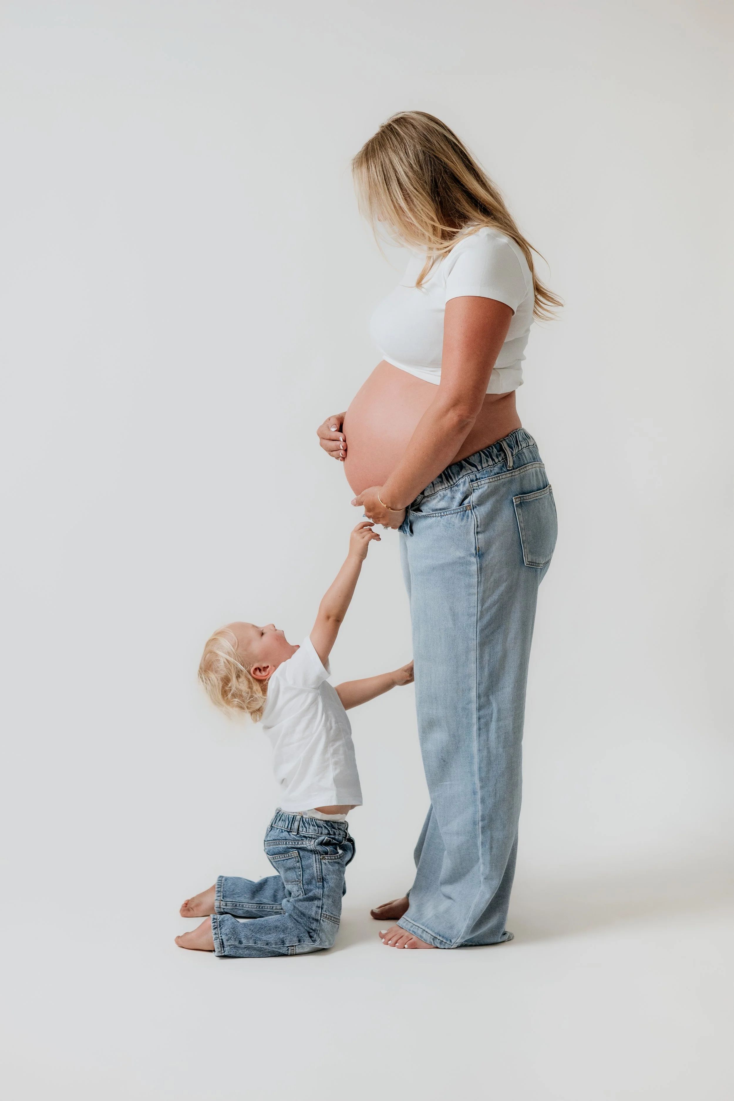 A pregnant woman in a white crop top and jeans holding her belly, with a young child kneeling and reaching up to her, all against a plain white background.