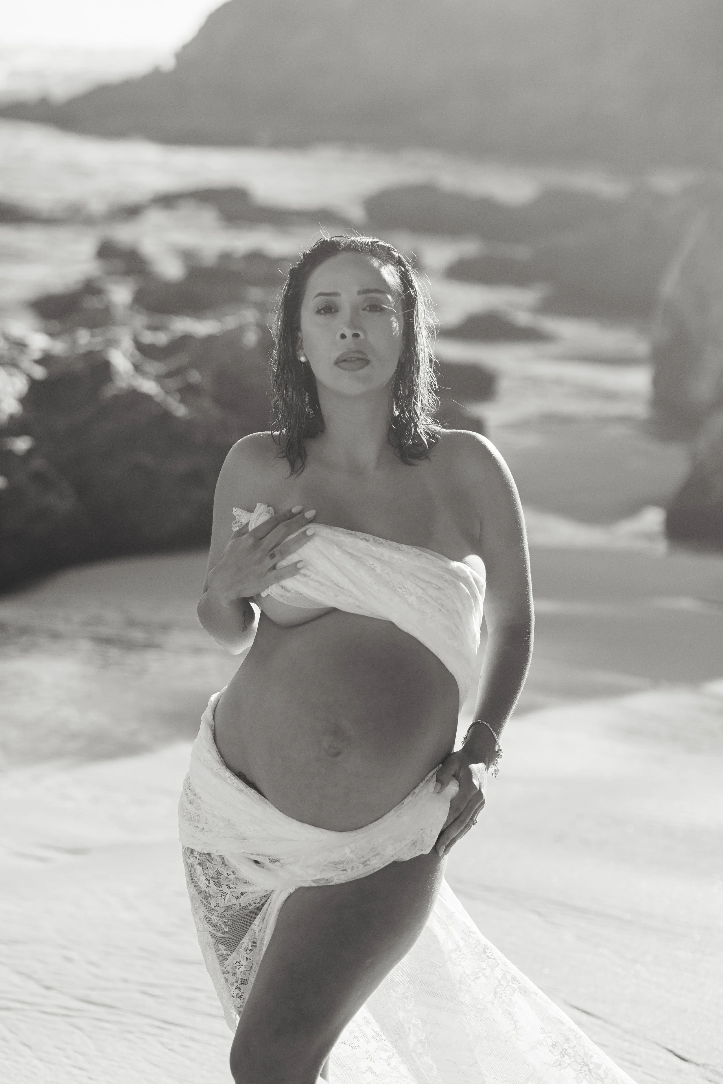 A woman with wet hair standing on a beach, holding a white cloth around her chest, with ocean waves and rocks in the background.
