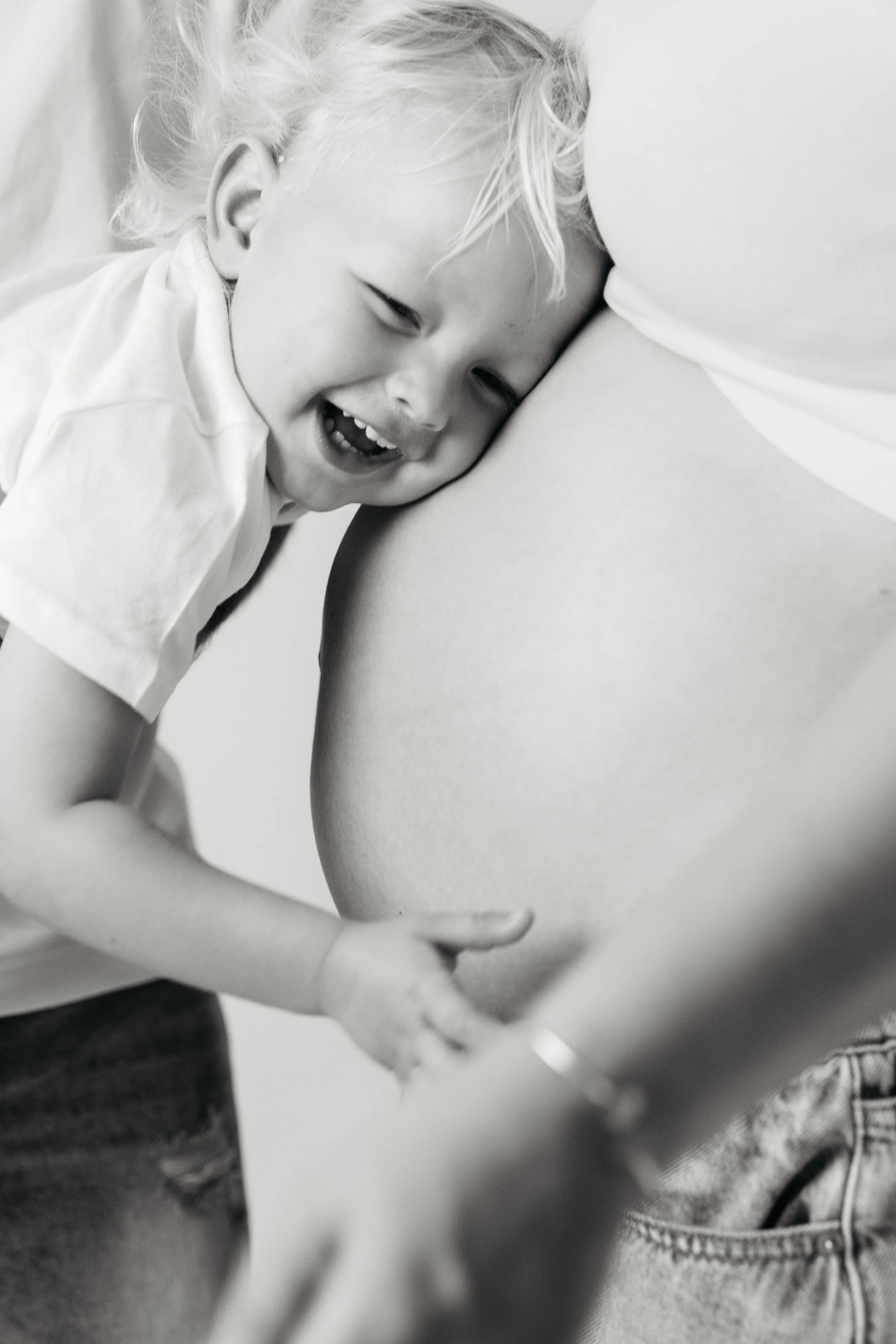 A young child with blonde hair smiling and laughing, leaning against an adult's arm, close-up in black and white.