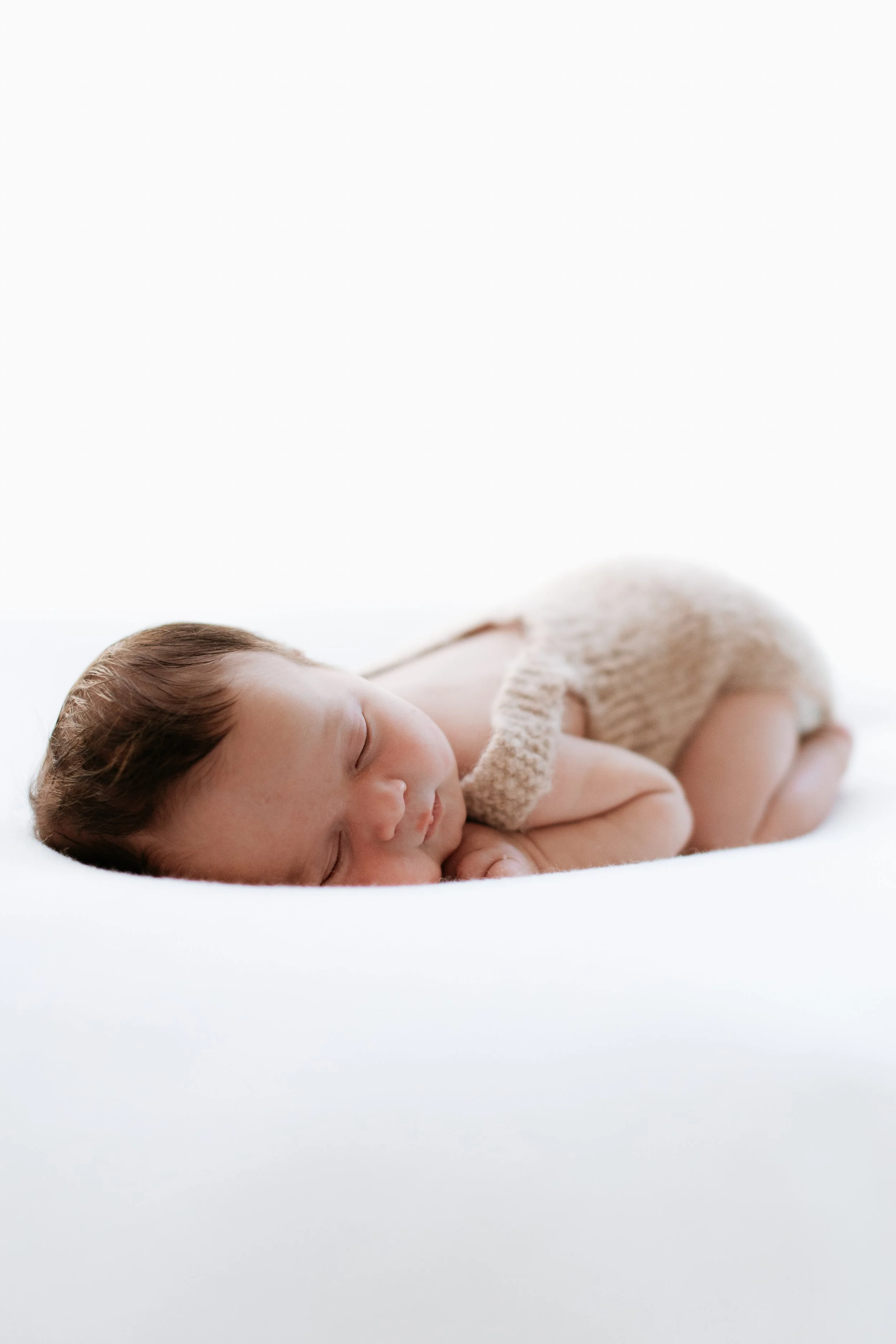 A sleeping infant with light skin, brown hair, and closed eyes, lying on a soft white surface, wearing a beige knitted outfit.