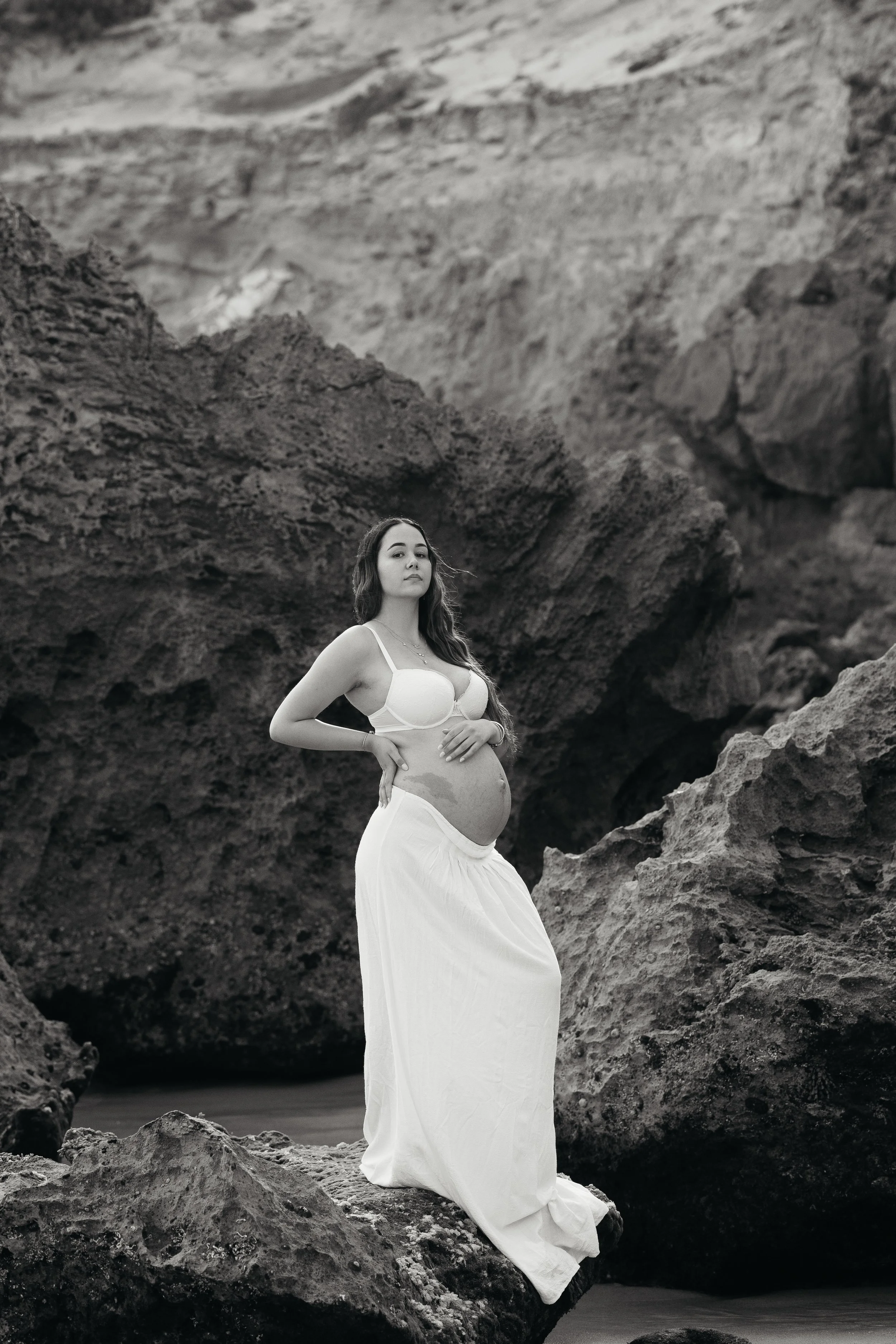 A pregnant woman in a white dress stands among rocks and volcanic formations on a beach.
