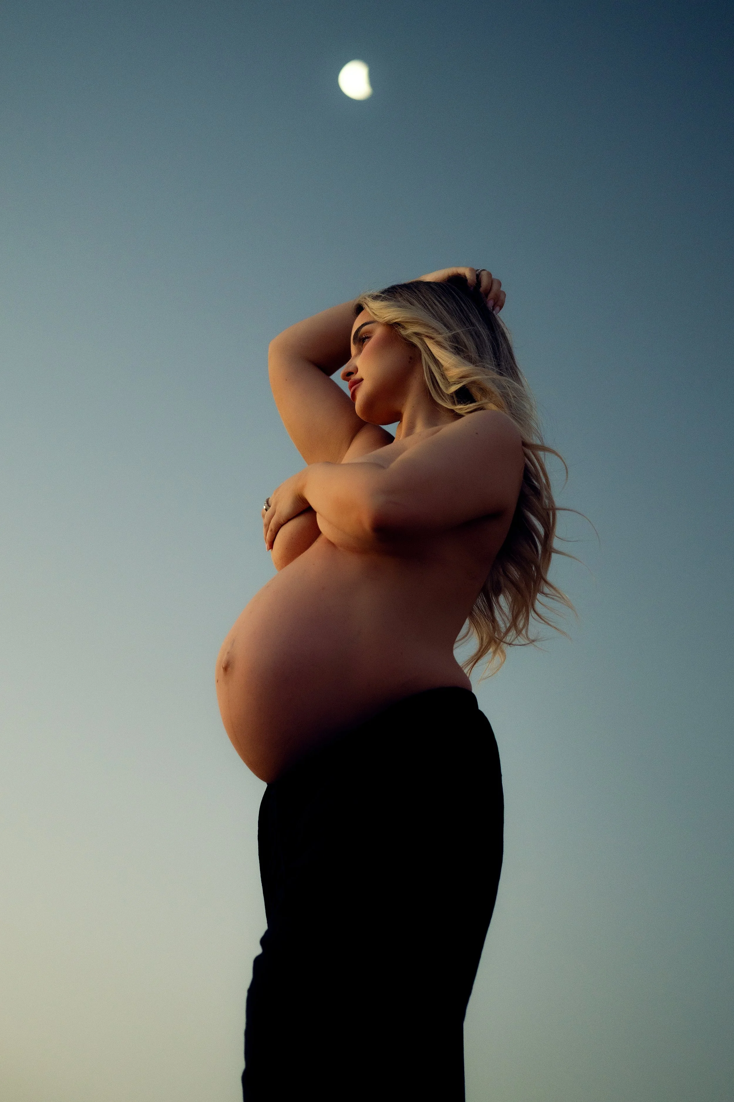 Pregnant woman standing outdoors during sunset, holding her belly and looking to the side with her eyes closed.