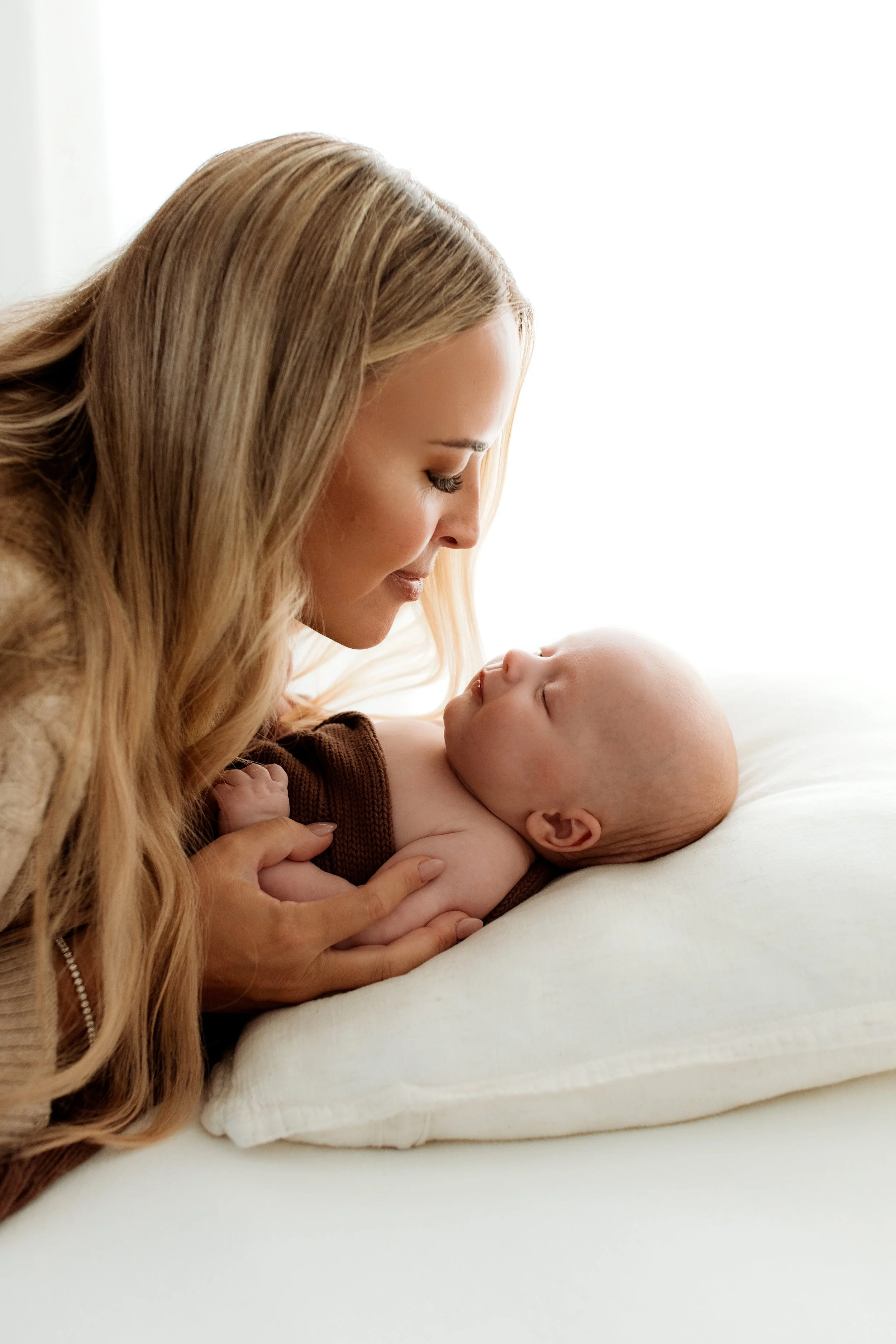 A woman with long blonde hair and a woman with a bald head are close to each other, smiling and looking into each other's eyes, with the woman on the bed.