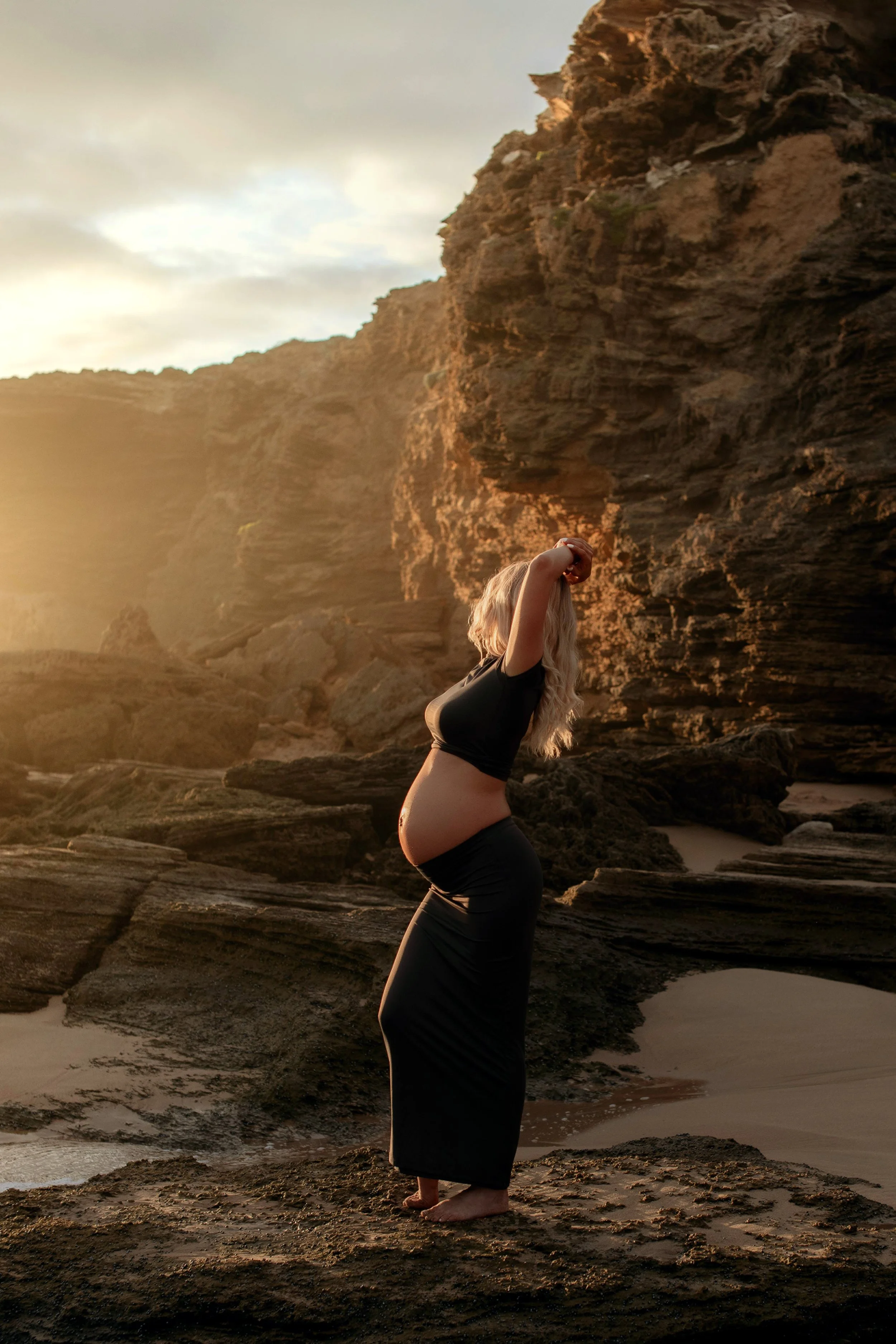 Luxury maternity photography in Melbourne featuring an expectant mother posing on a rocky beach at golden hour.
