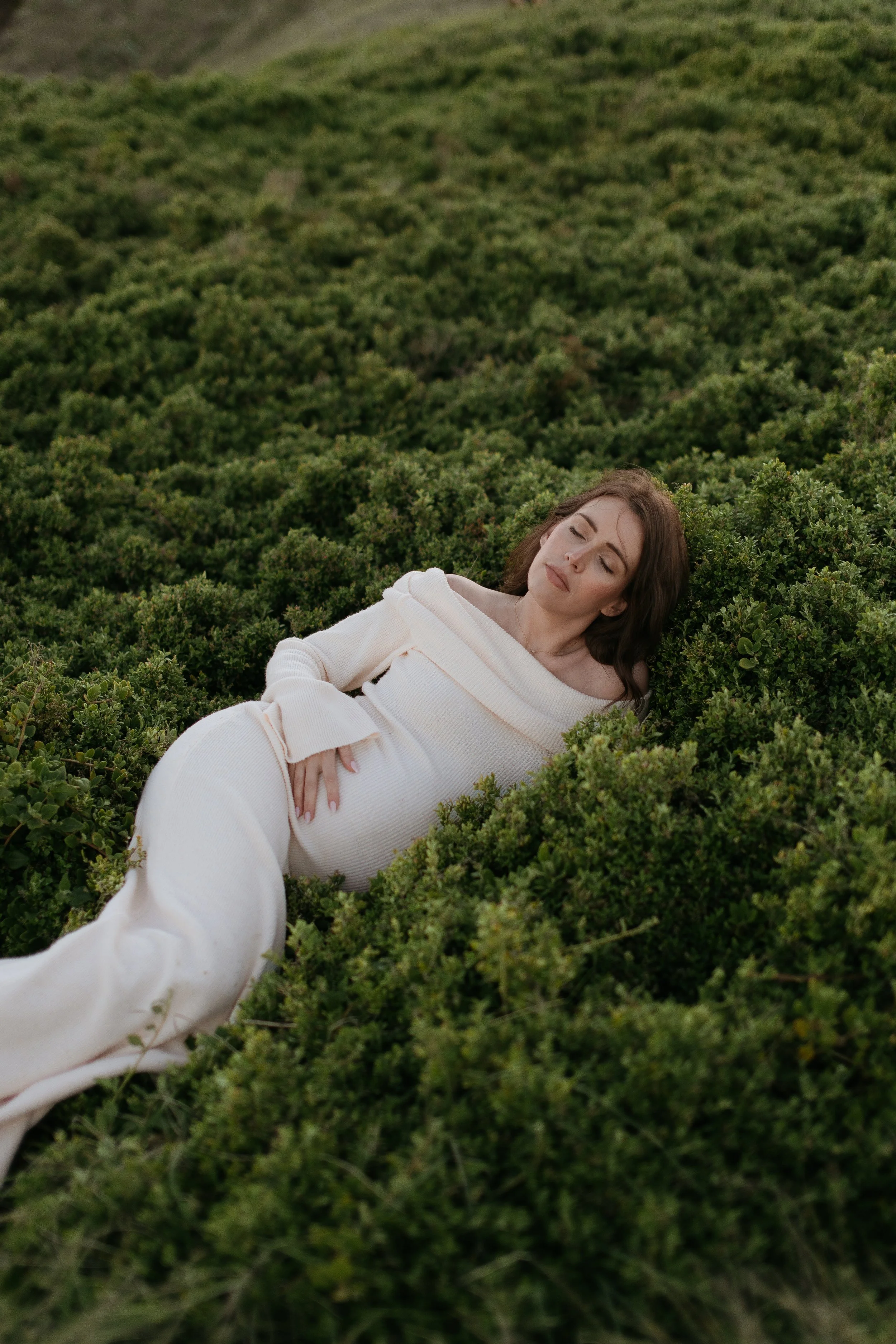 A woman in a white outfit lying on green shrubbery with her eyes closed.