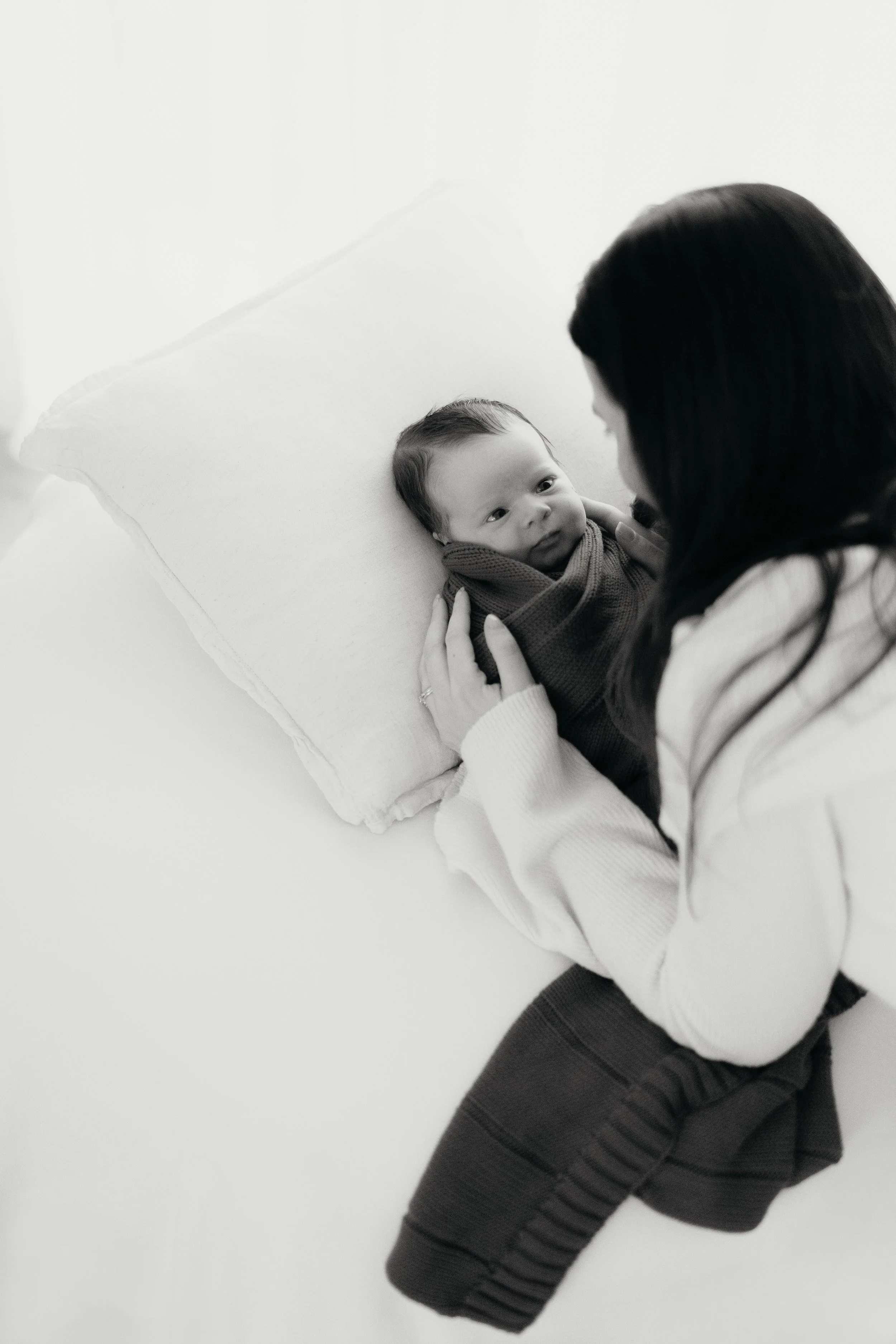 A woman holding a baby in her arms while lying on a white bed or couch, with a large white pillow behind the baby. The woman has long dark hair and is wearing a light-colored sweater, while the baby is wrapped in a blanket and looking up at her.