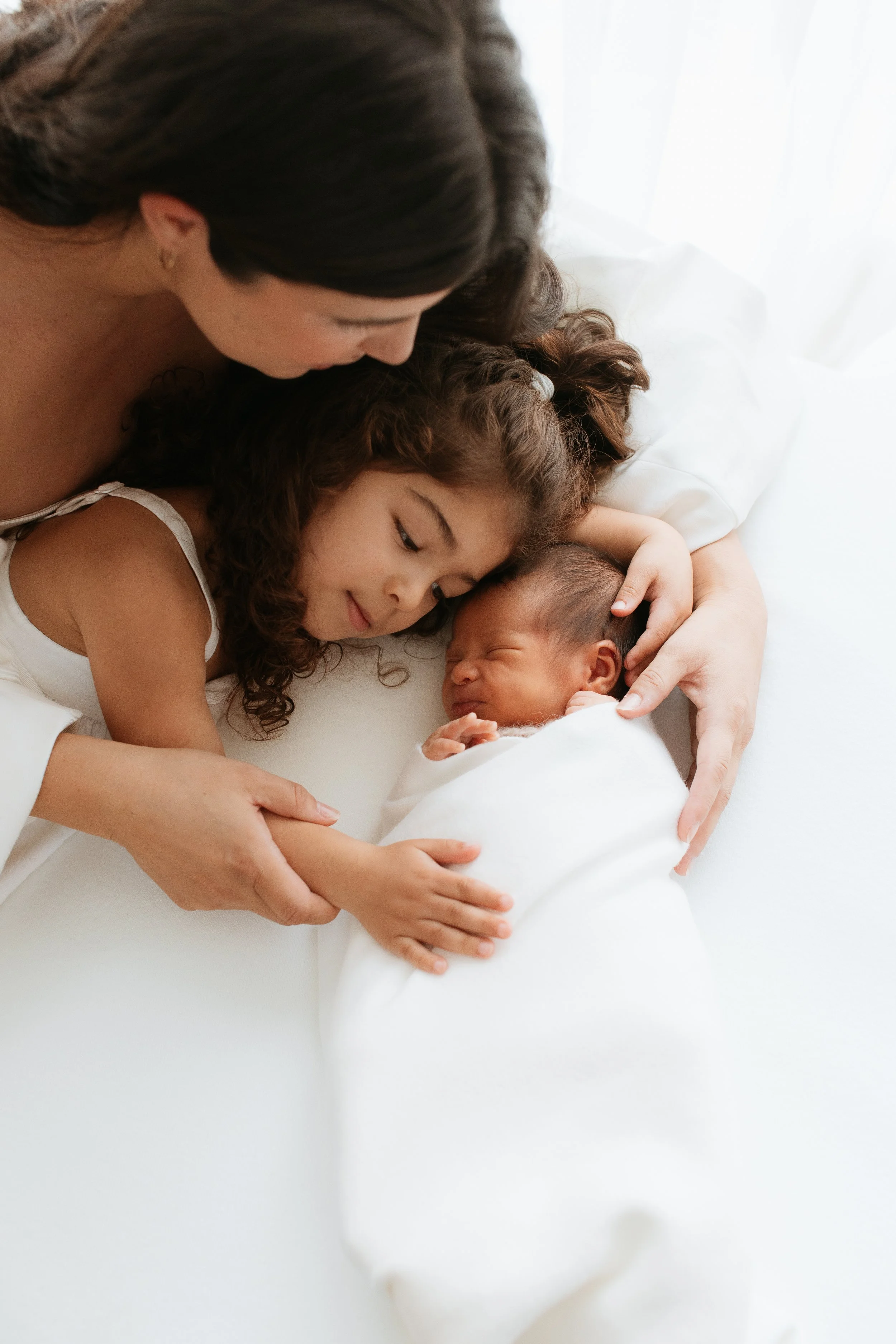 Natural newborn family photography in Melbourne featuring a mother cuddling her newborn baby with an older sibling in a bright, timeless portrait.