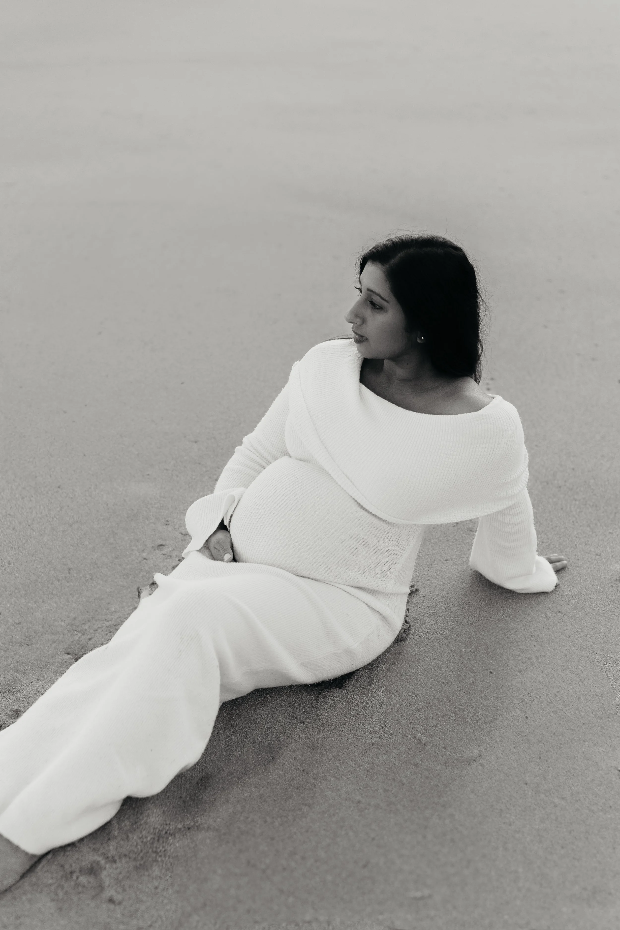 Editorial maternity photography in Melbourne featuring an expectant mother in an elegant white dress posing on the beach in a timeless black and white portrait.