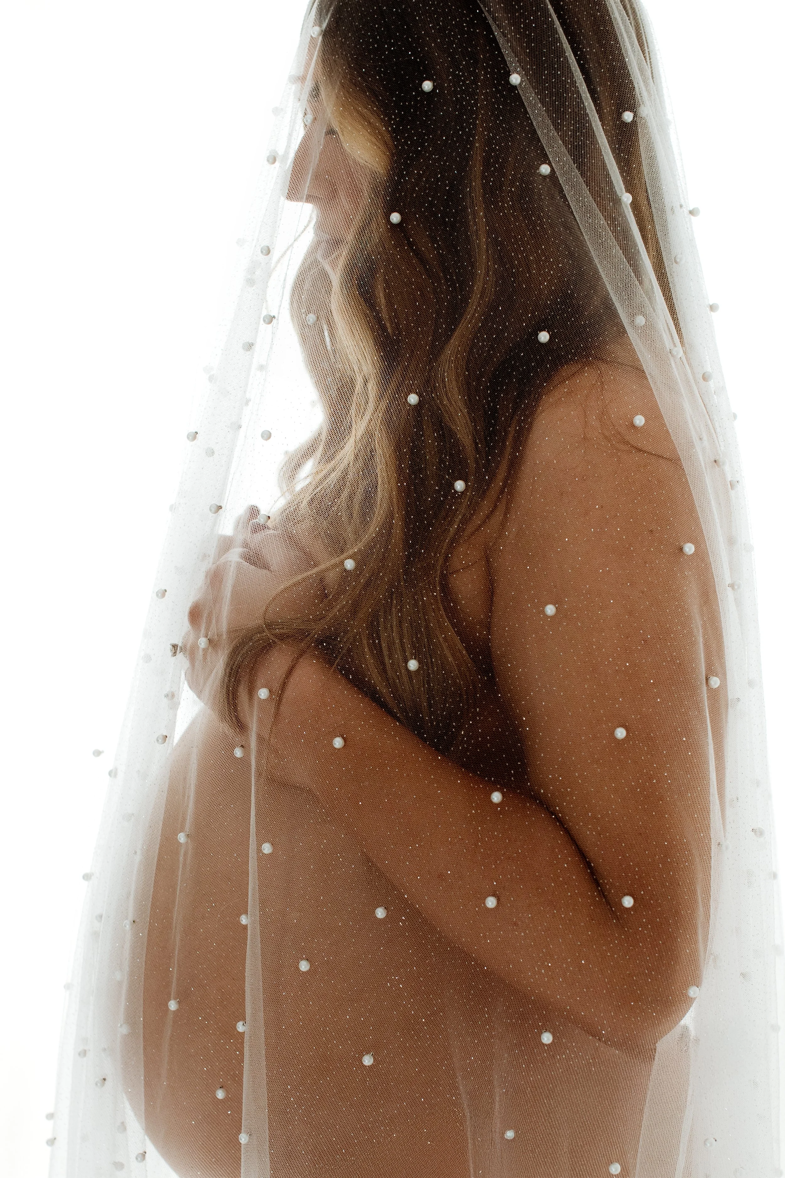 A woman with long, wavy hair is behind a sheer veil adorned with small pearls, covering her face and upper body.