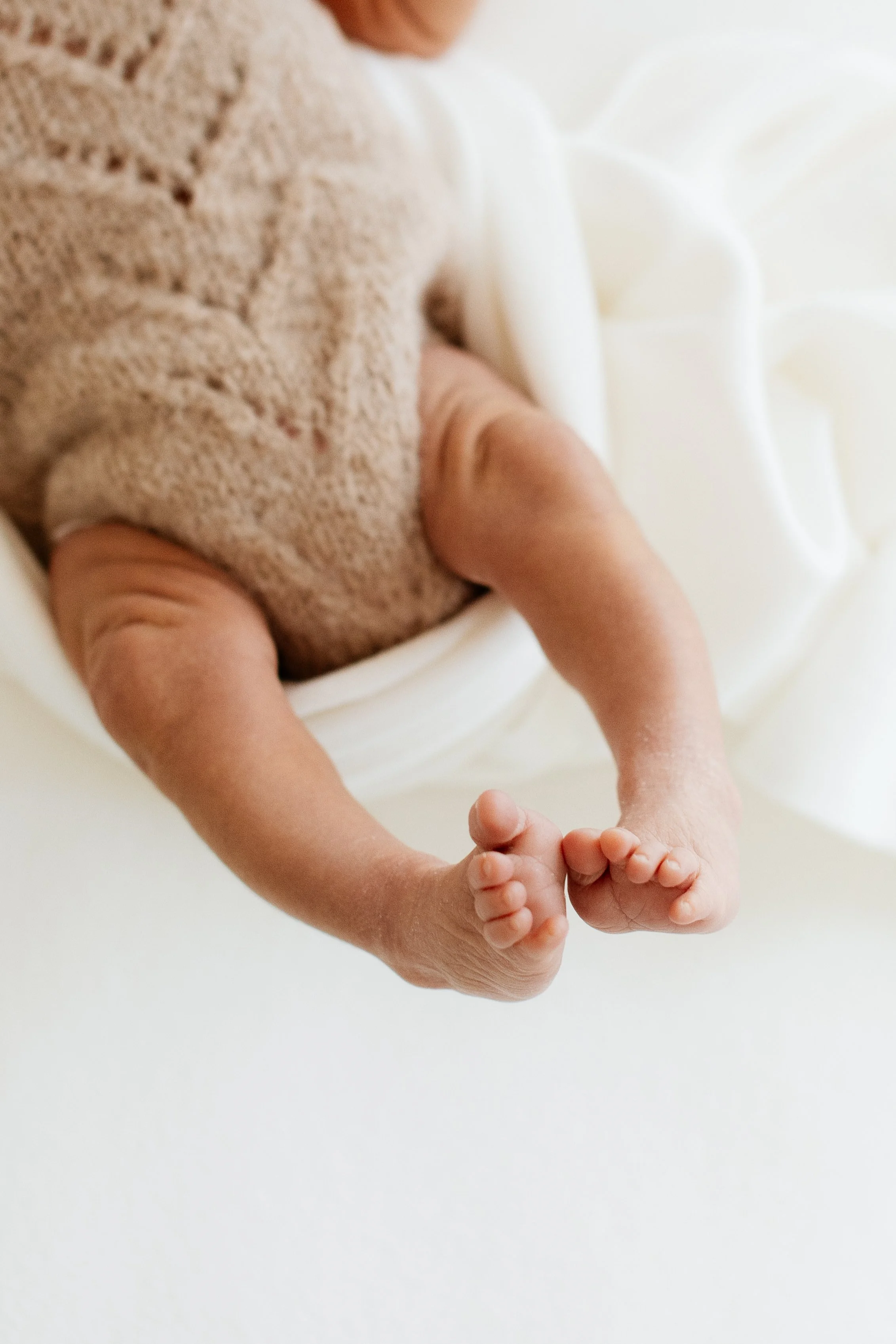 Close-up of a newborn baby lying on a white surface, wearing a beige knitted outfit, holding an adult's finger, with legs crossed and tiny toes visible.