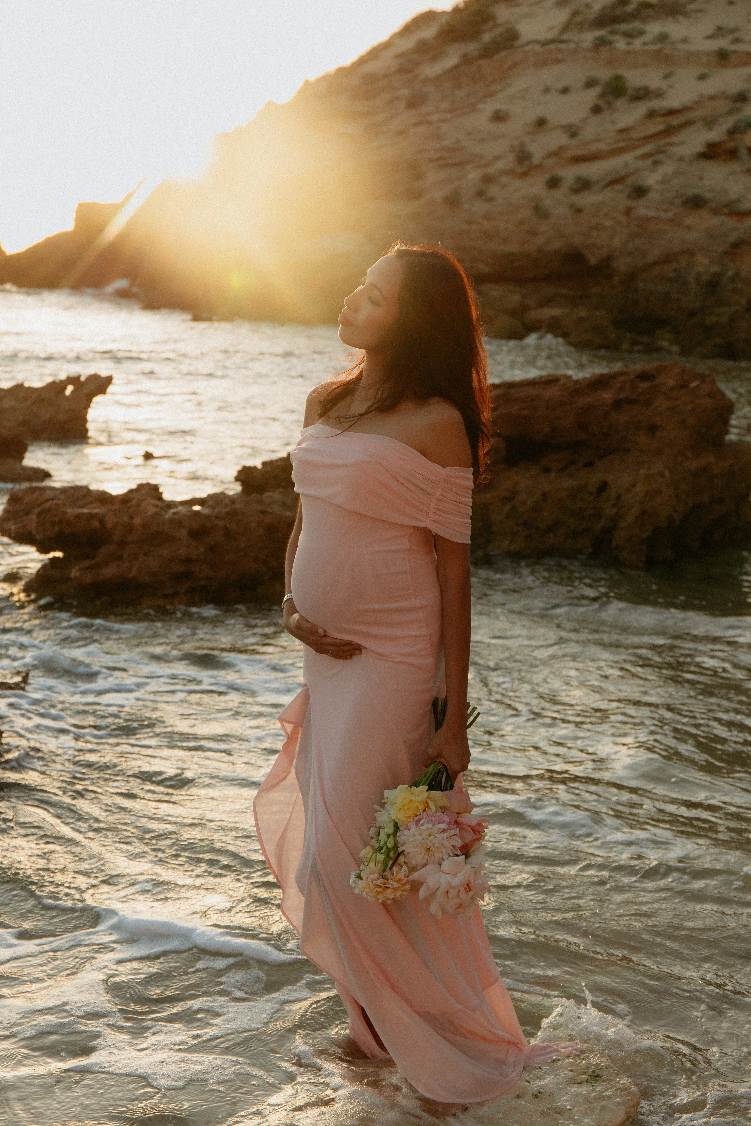 Luxury beach maternity photography in Melbourne capturing an expectant mother standing in the ocean at sunset holding a bouquet of flowers.
