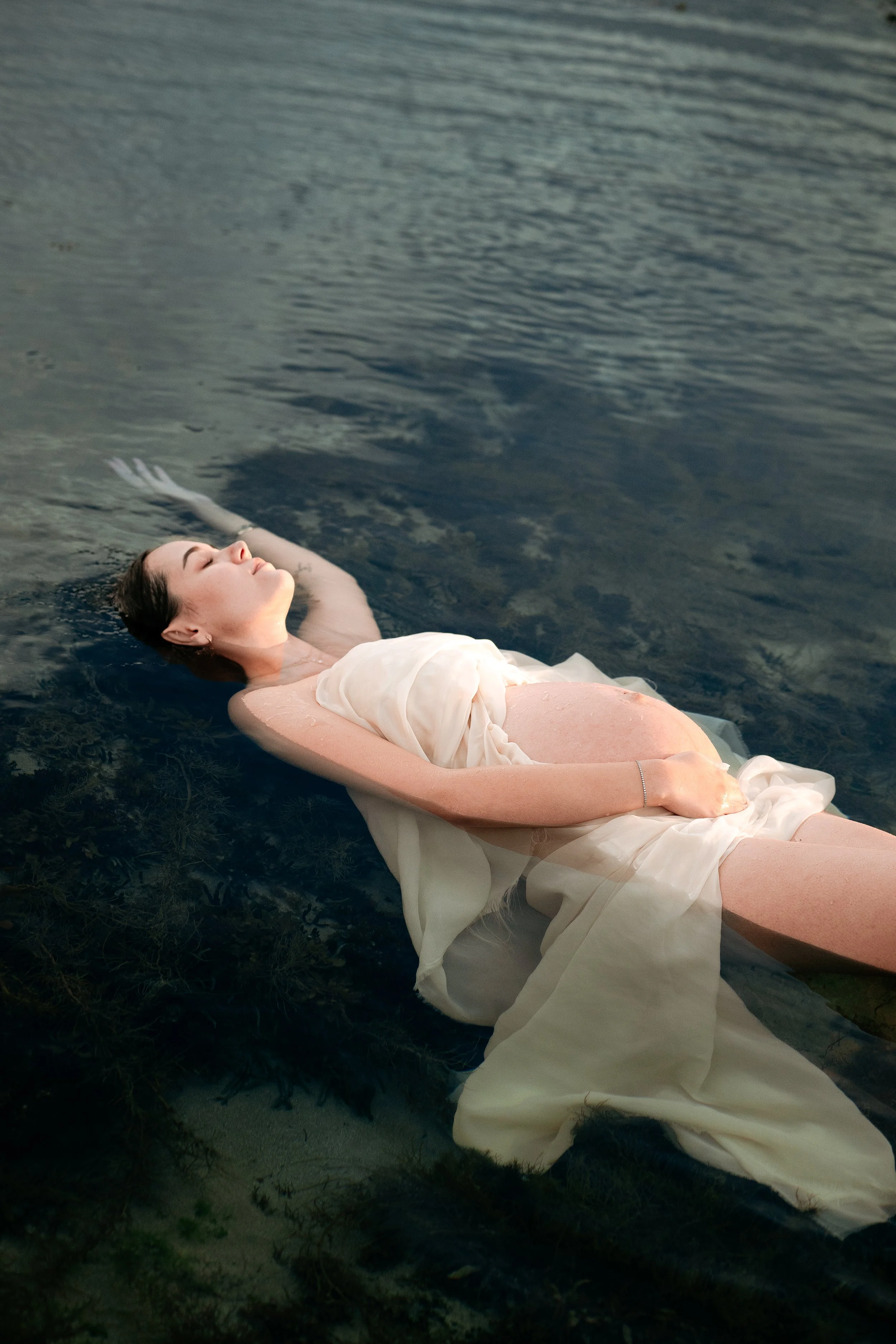 A young woman with closed eyes floating on water, wearing a white dress, surrounded by dark green seaweed under a cloudy sky.