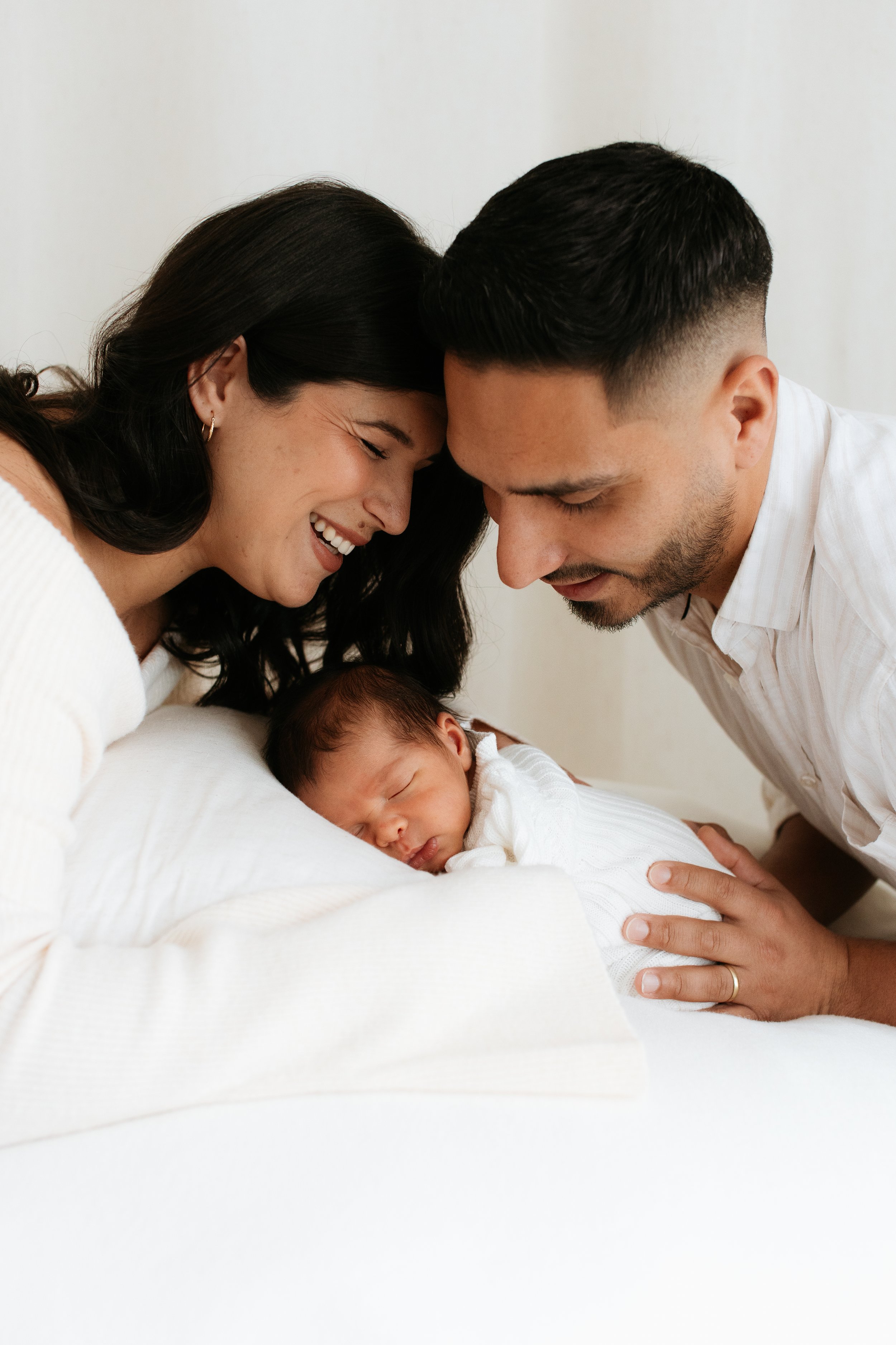 A happy woman and man leaning over, touching foreheads and smiling at a sleeping newborn baby, all dressed in white, in a minimalistic setting.