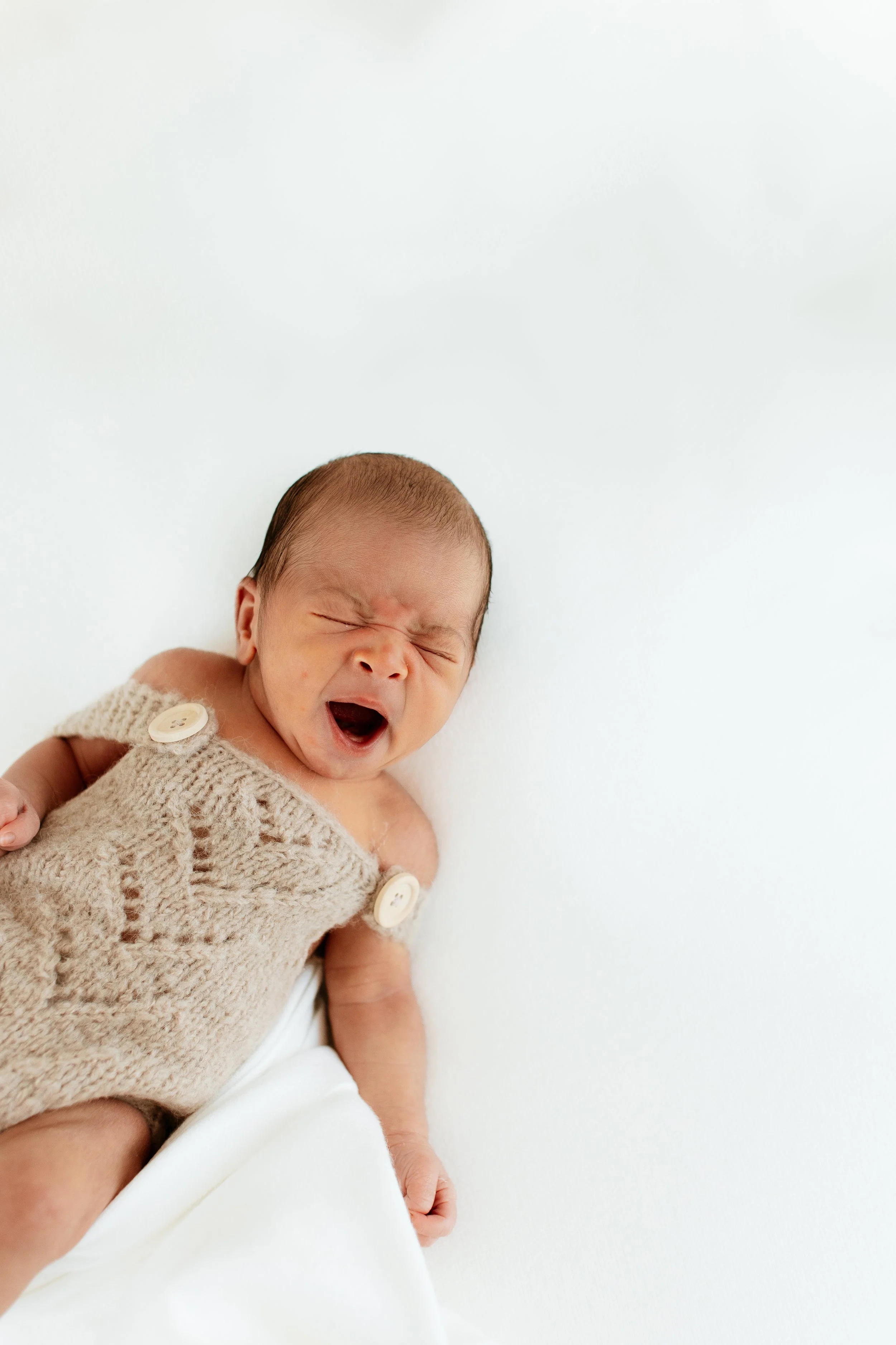 A newborn baby with a scrunched face and open mouth, wrapped in a hand-knit beige vest with large buttons, on a white surface.