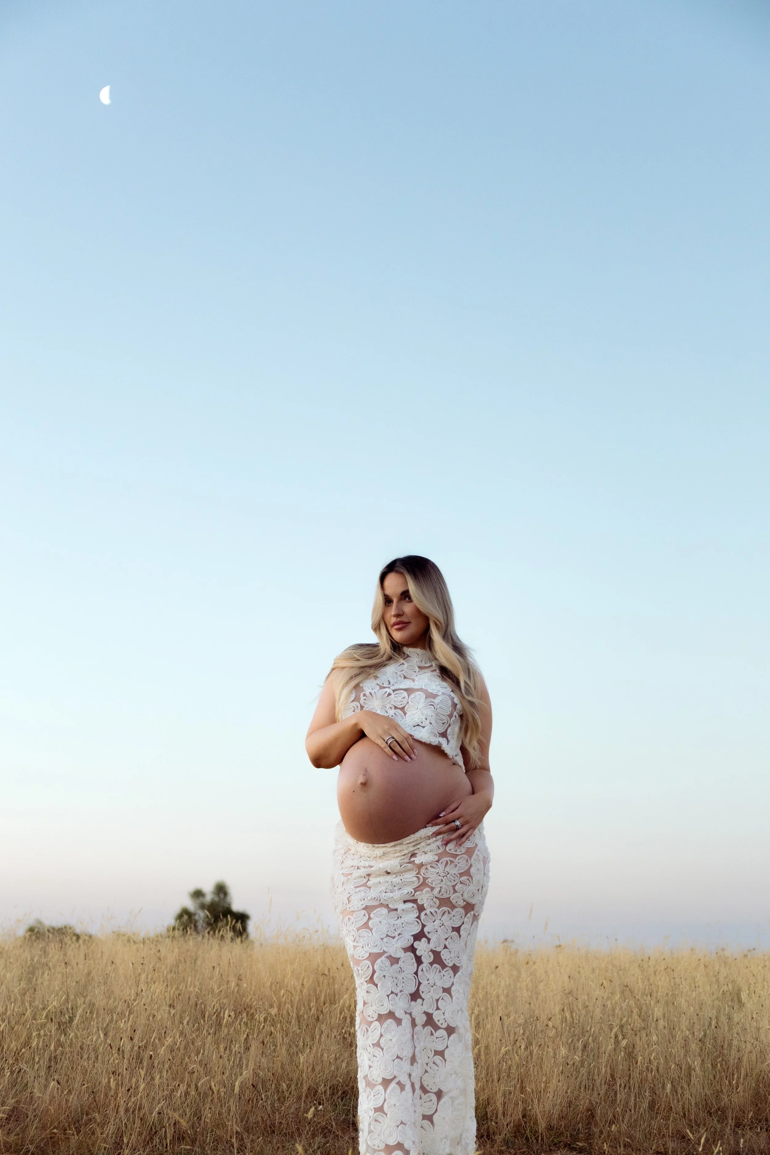A pregnant woman in a white lace dress standing in a field of tall grass during sunset, with a clear sky and moon in the background.