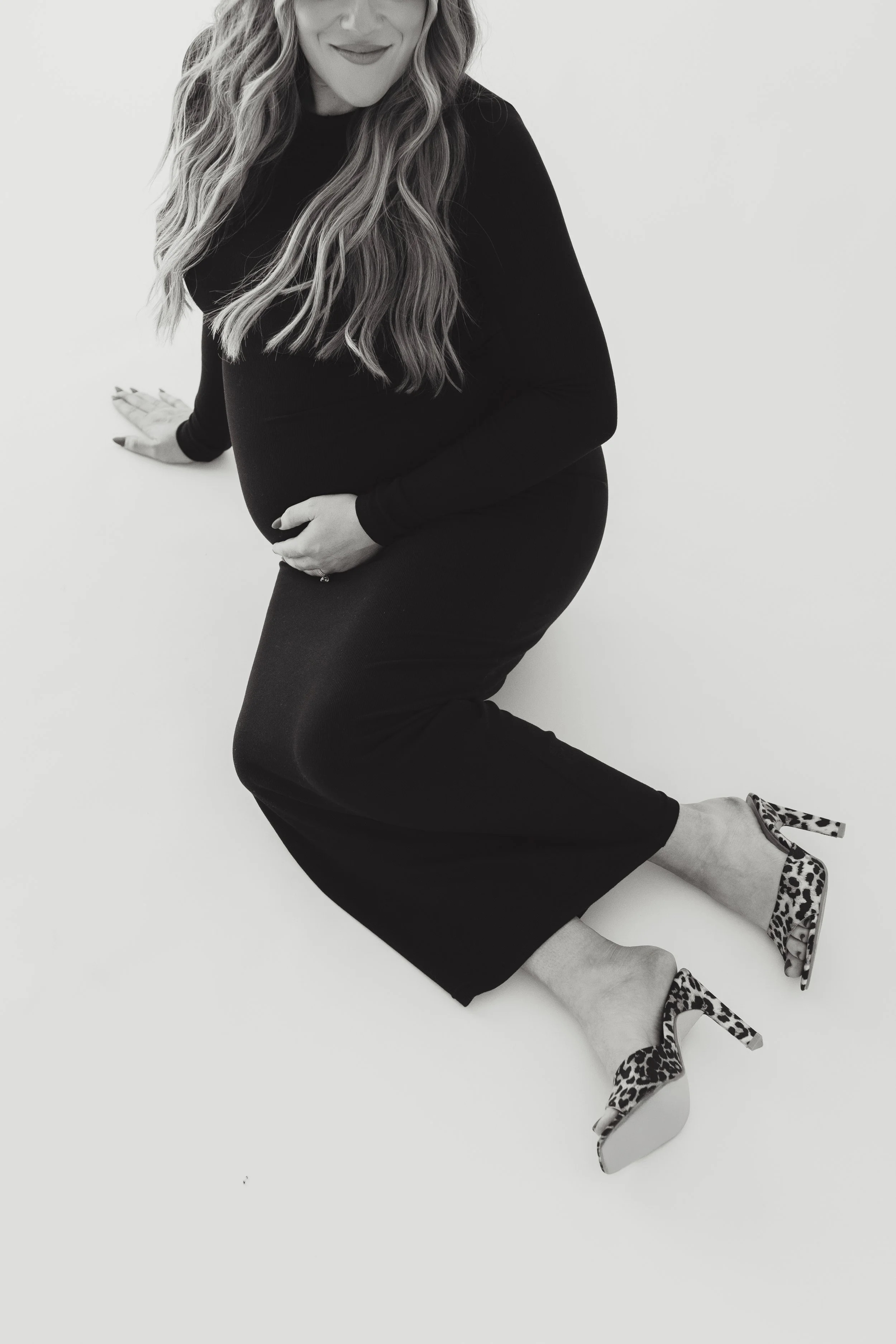 Black and white photo of a woman in a black dress, lying on her side on a white surface, wearing leopard print high heels, smiling.