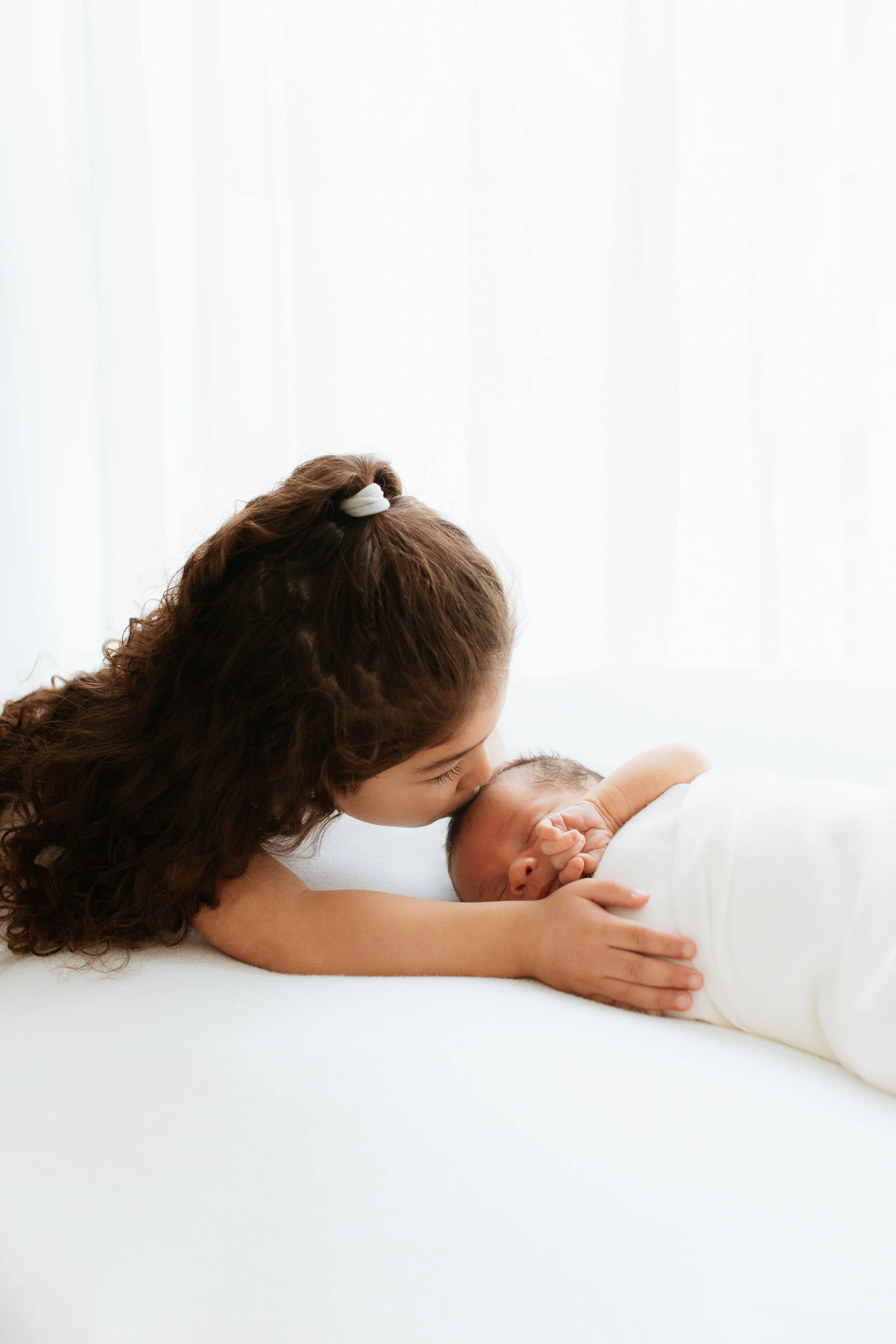 A woman with long, curly brown hair and a white hair clip kisses a sleeping newborn baby wrapped in a white blanket.