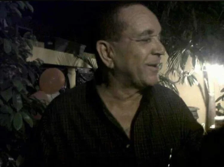 A smiling man with short hair and glasses, wearing a dark checkered shirt, standing outdoors at night with plants and string lights in the background.