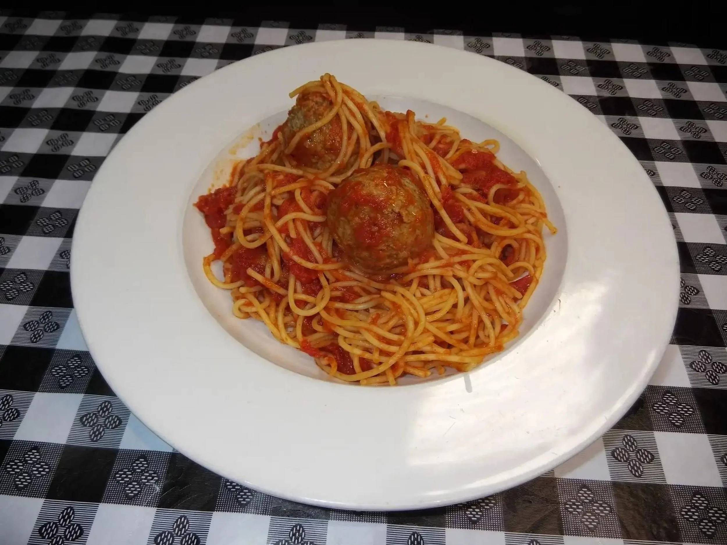 A plate of spaghetti with meatballs in tomato sauce on a white dish, placed on a black and white checkered tablecloth.
