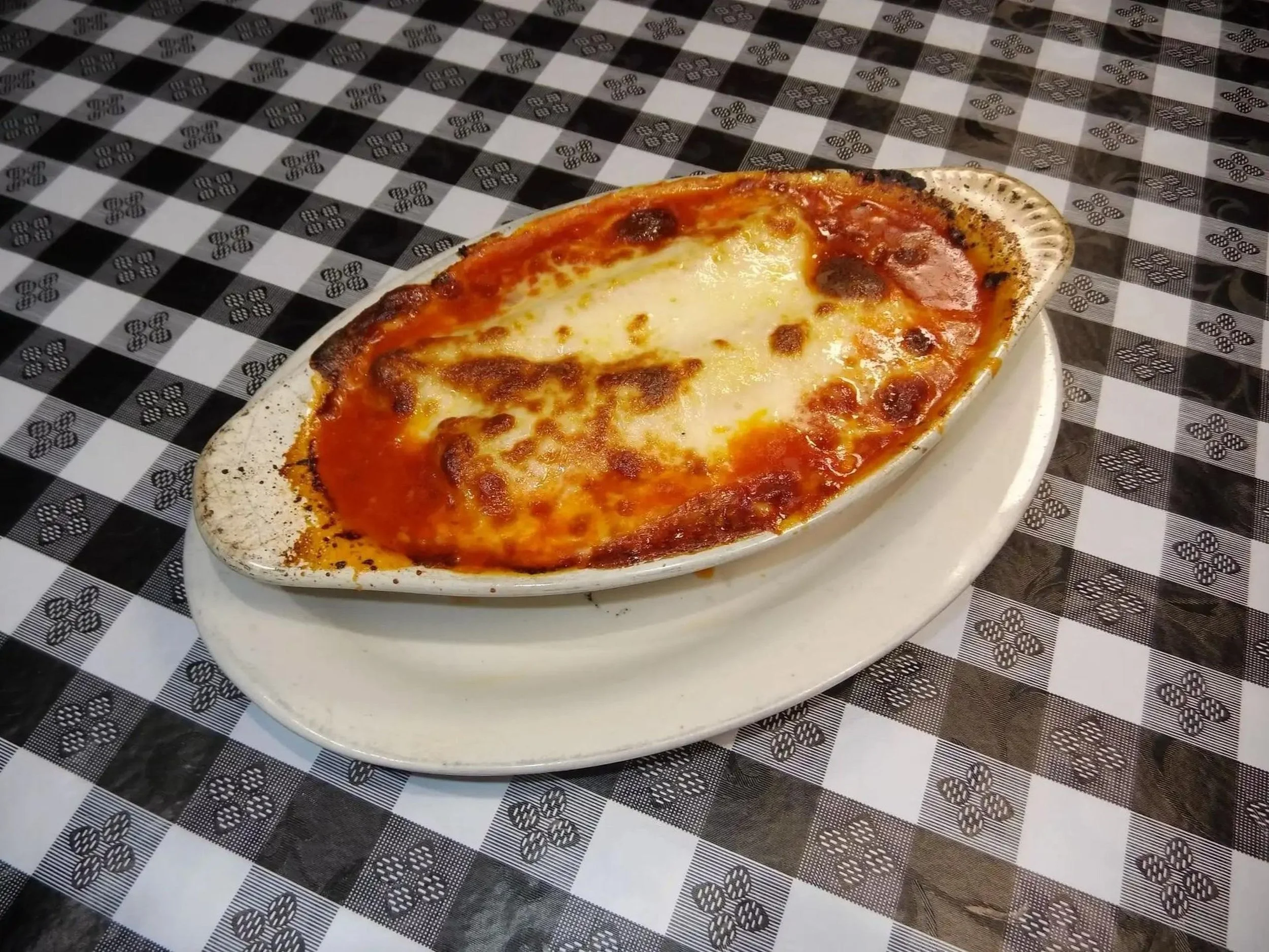 Baked pizza with cheese and tomato sauce on a white oval dish placed on a black and white checkered tablecloth.