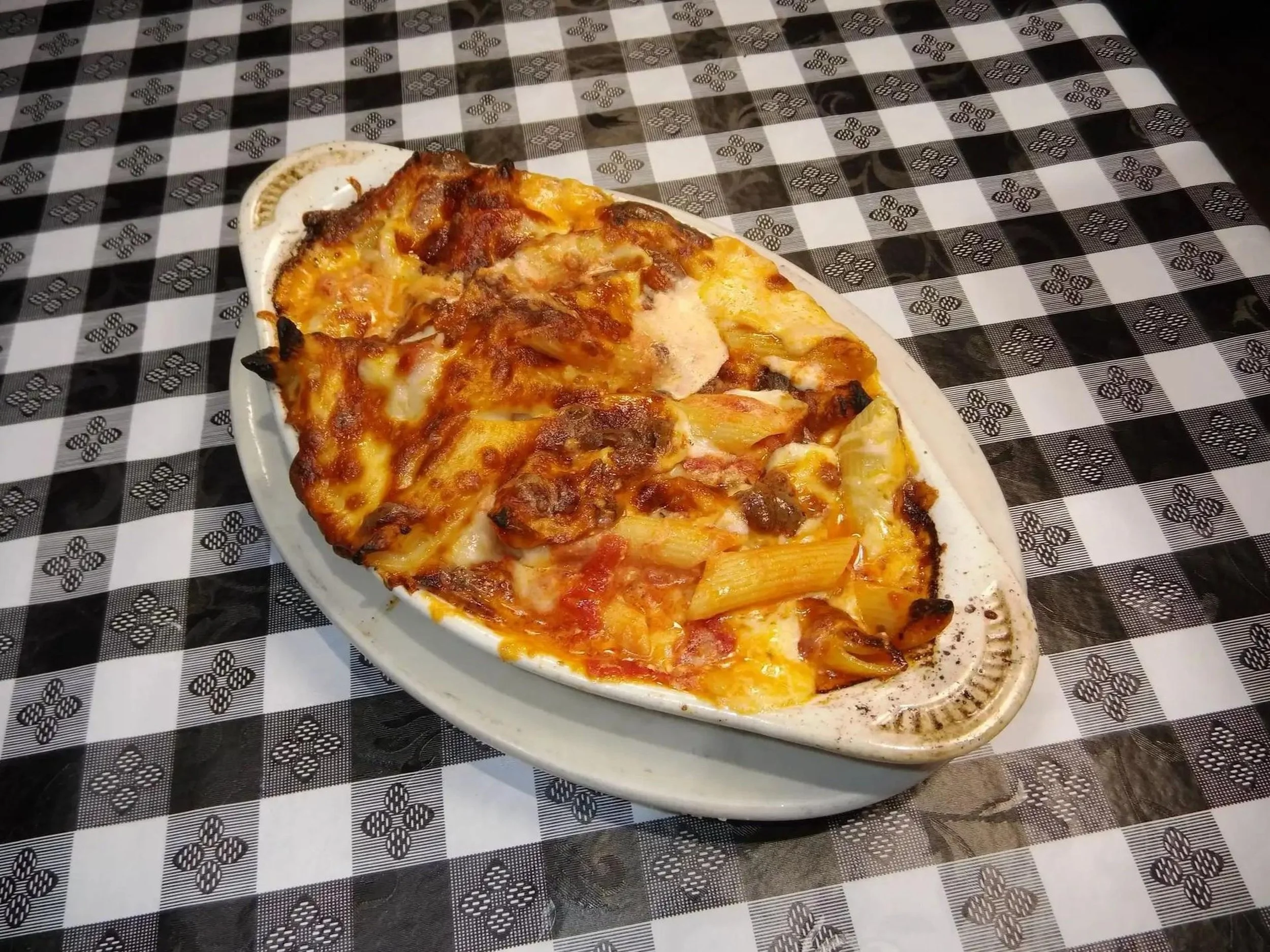 Baked pasta with melted cheese and tomato sauce served on a white oval dish on a black and white checkered tablecloth.