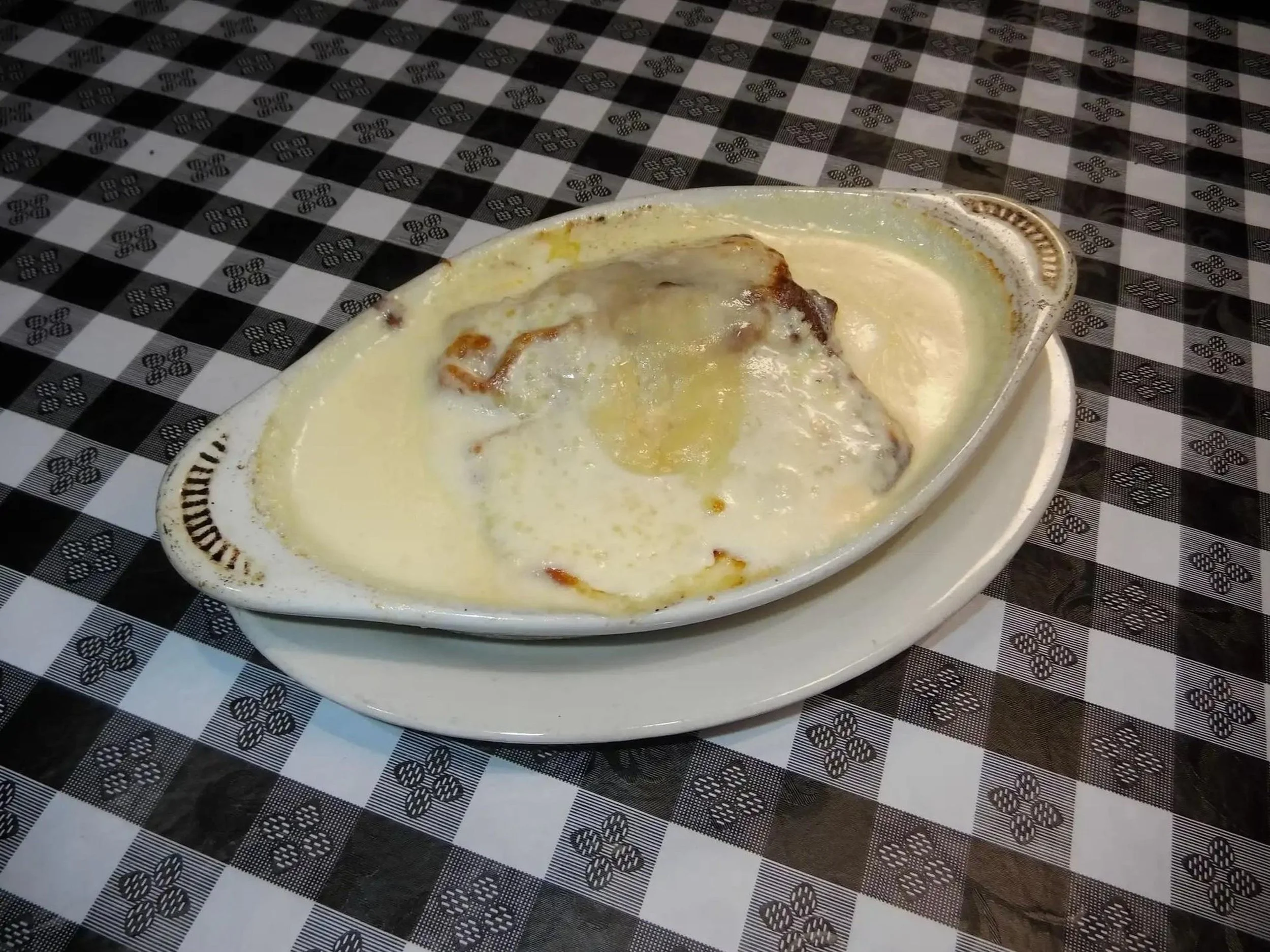 A dish of baked casserole with a creamy, cheesy topping, served in an oval ceramic dish on a black and white checkered tablecloth.