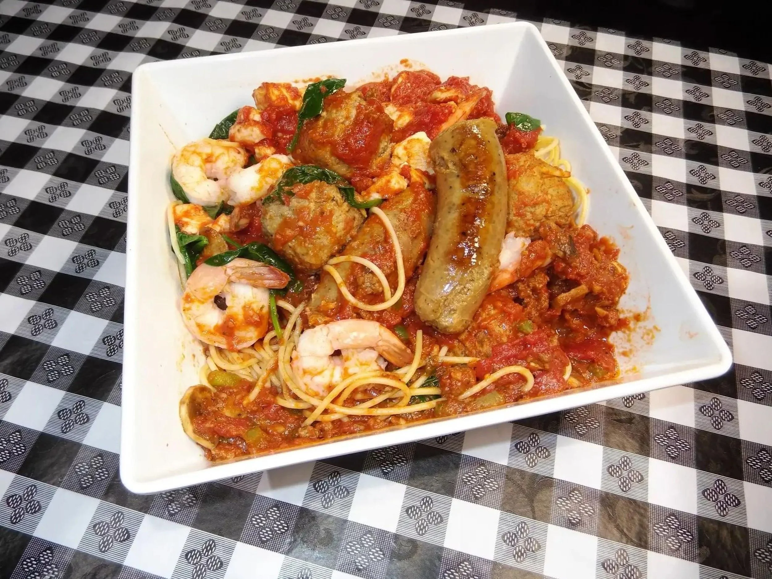 A white rectangular plate with spaghetti, shrimp, meatballs, sausage, and tomato sauce, garnished with spinach leaves, on a black-and-white checkered tablecloth.